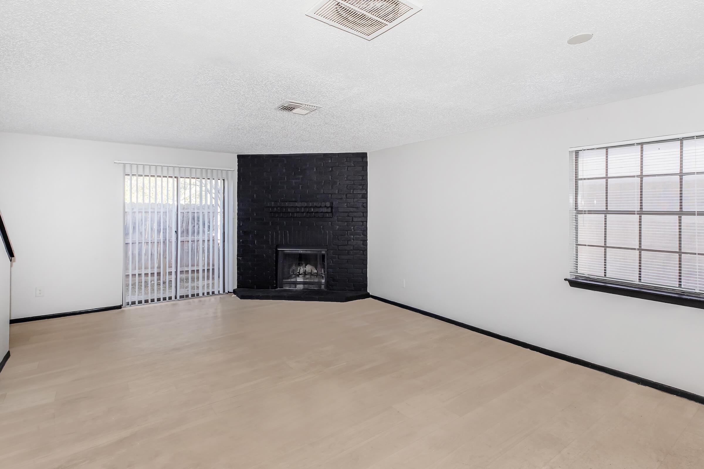 A spacious living room featuring light-colored walls, a black-brick fireplace, large windows with blinds, and access to a small outdoor area through sliding glass doors. The flooring is light wood, creating a bright and open atmosphere.