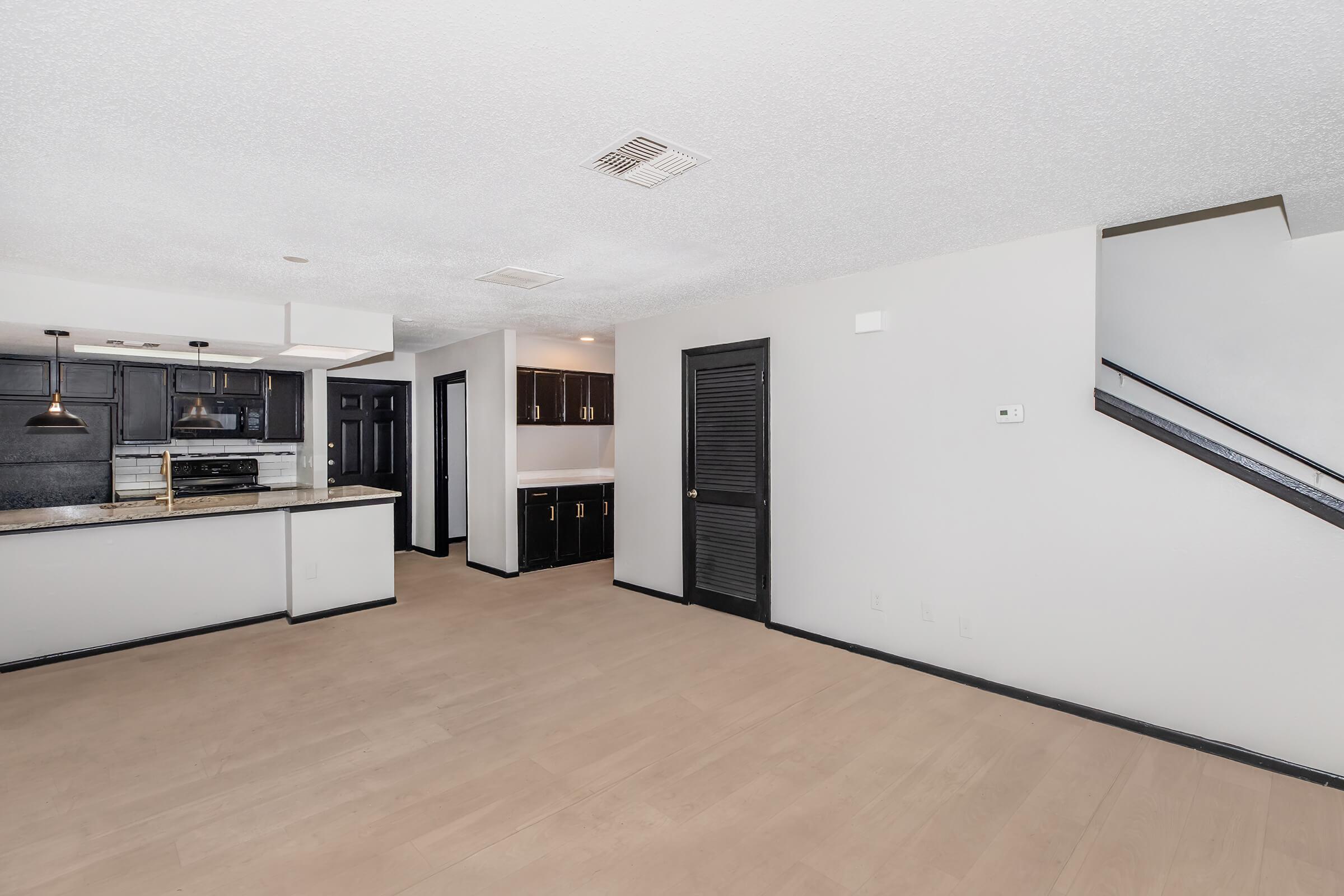 Spacious interior of a modern living area featuring light-colored flooring, black kitchen cabinetry, and an open layout. The room has a staircase in the corner, with walls painted in a neutral color. Bright overhead lighting and a large window contribute to a well-lit ambiance.