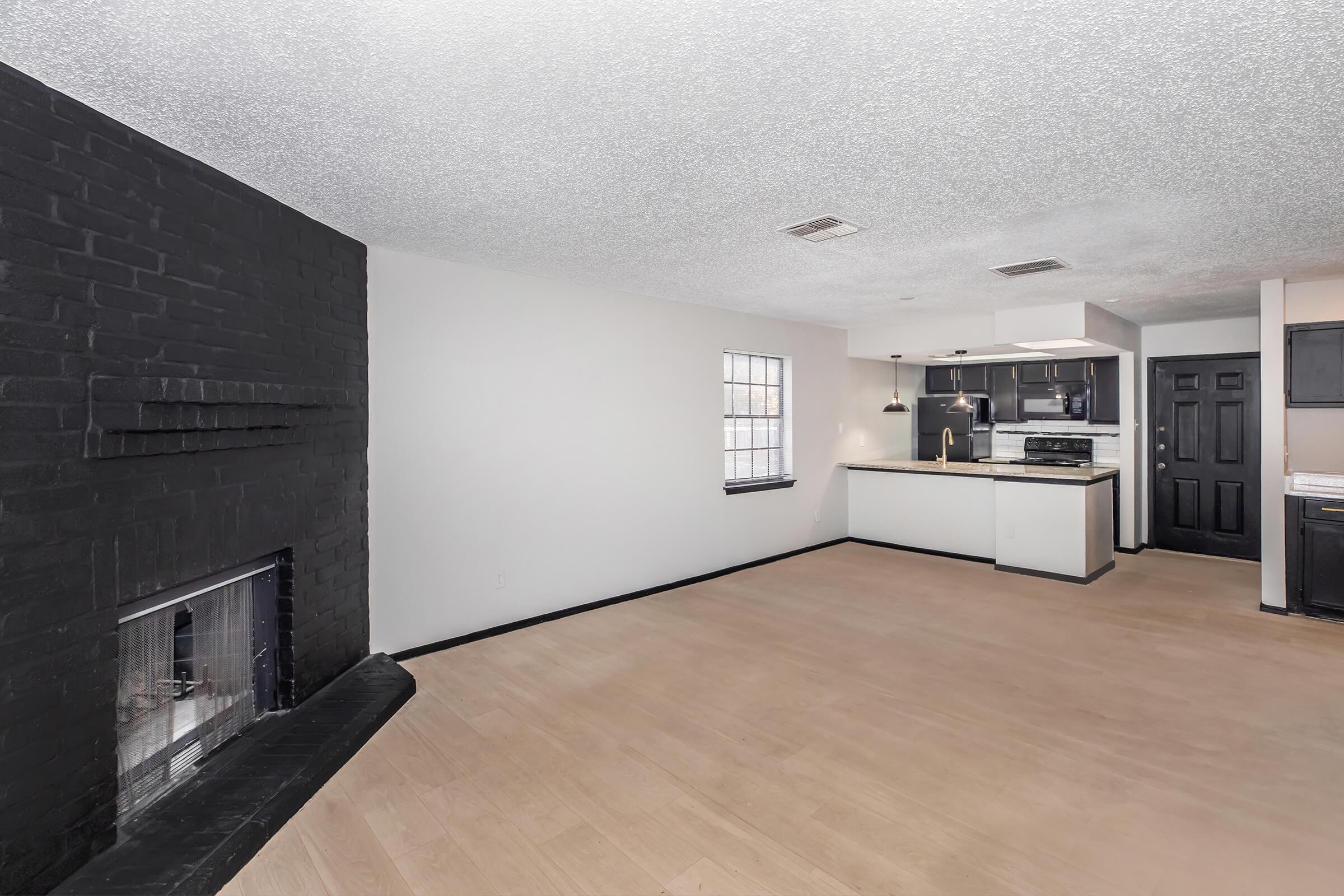 A spacious living area featuring a black fireplace, light-colored walls, and wooden flooring. The room is open to a kitchen area, equipped with modern appliances and a small bar counter. Large windows provide natural light, and there's a minimalist aesthetic with neutral colors and simple design.