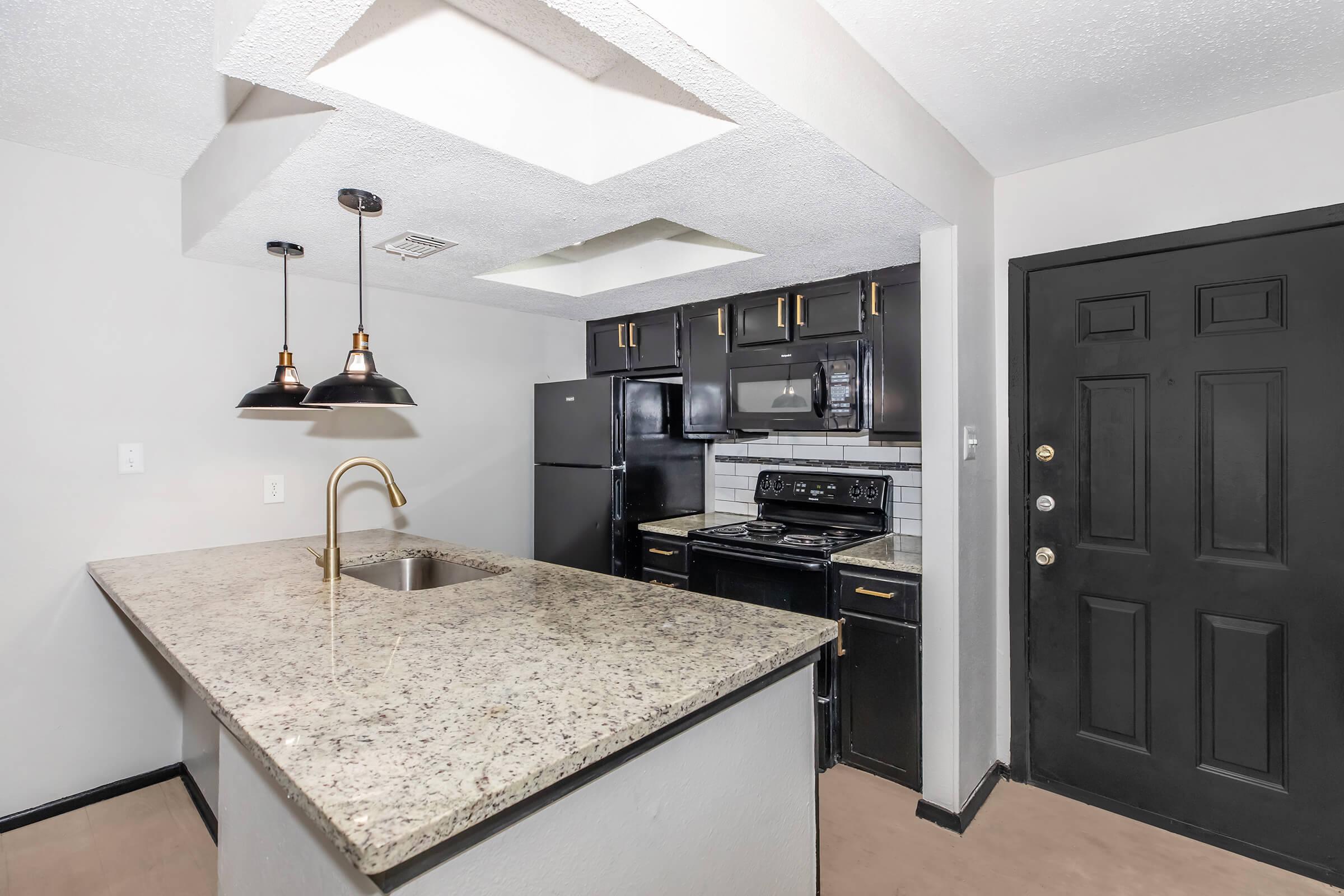 A modern kitchen featuring dark cabinetry, granite countertops, and stainless steel appliances. The kitchen island includes a sink and pendant lighting. A black refrigerator and stove are visible, along with a wall-mounted microwave. The entry door is black, complementing the overall design.
