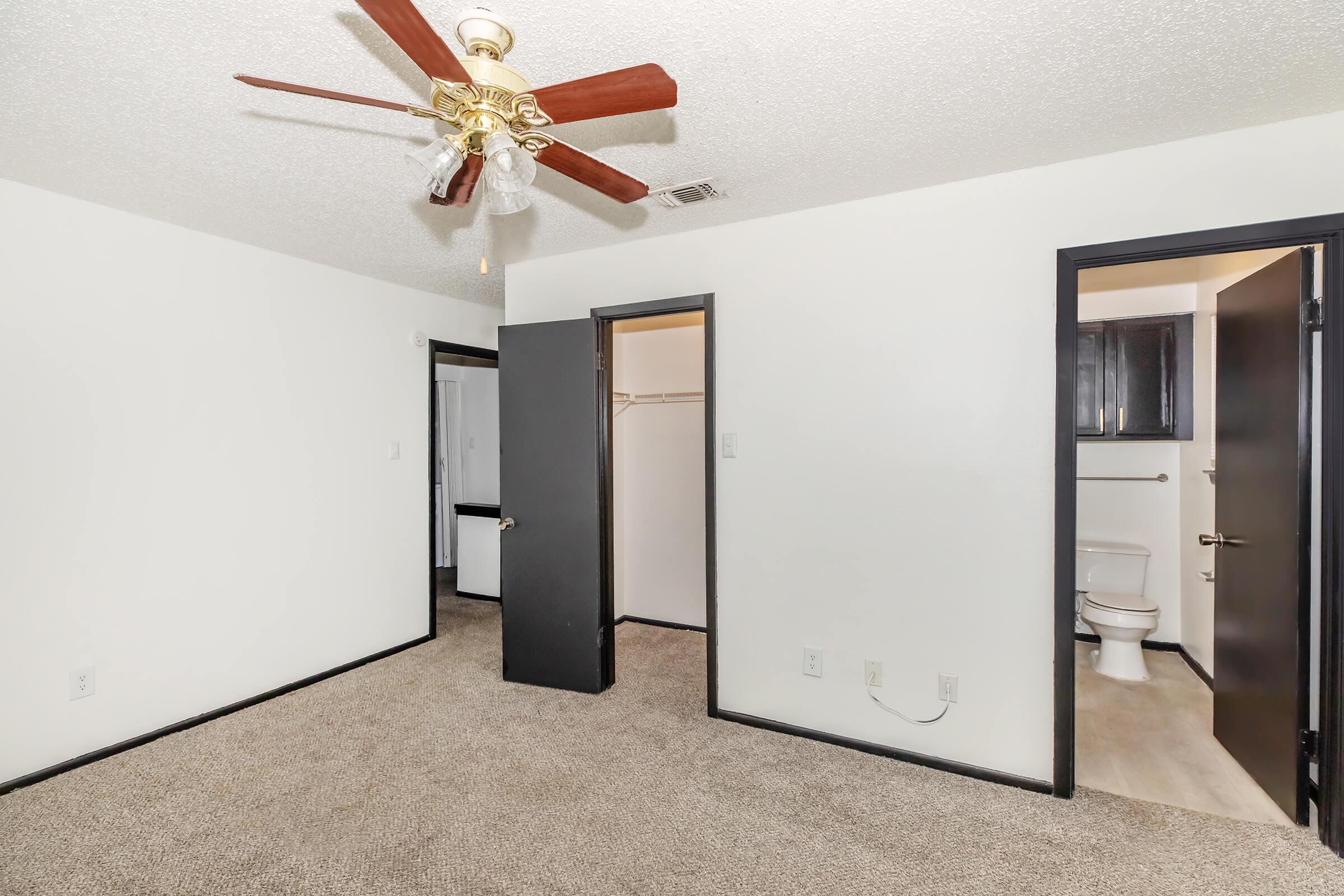 A well-lit, unfurnished bedroom featuring beige carpeting, a ceiling fan with wooden blades, and two doors leading to a closet and bathroom. The walls are painted white, and the doors and trim are black. The space is clean and ready for personal decoration.