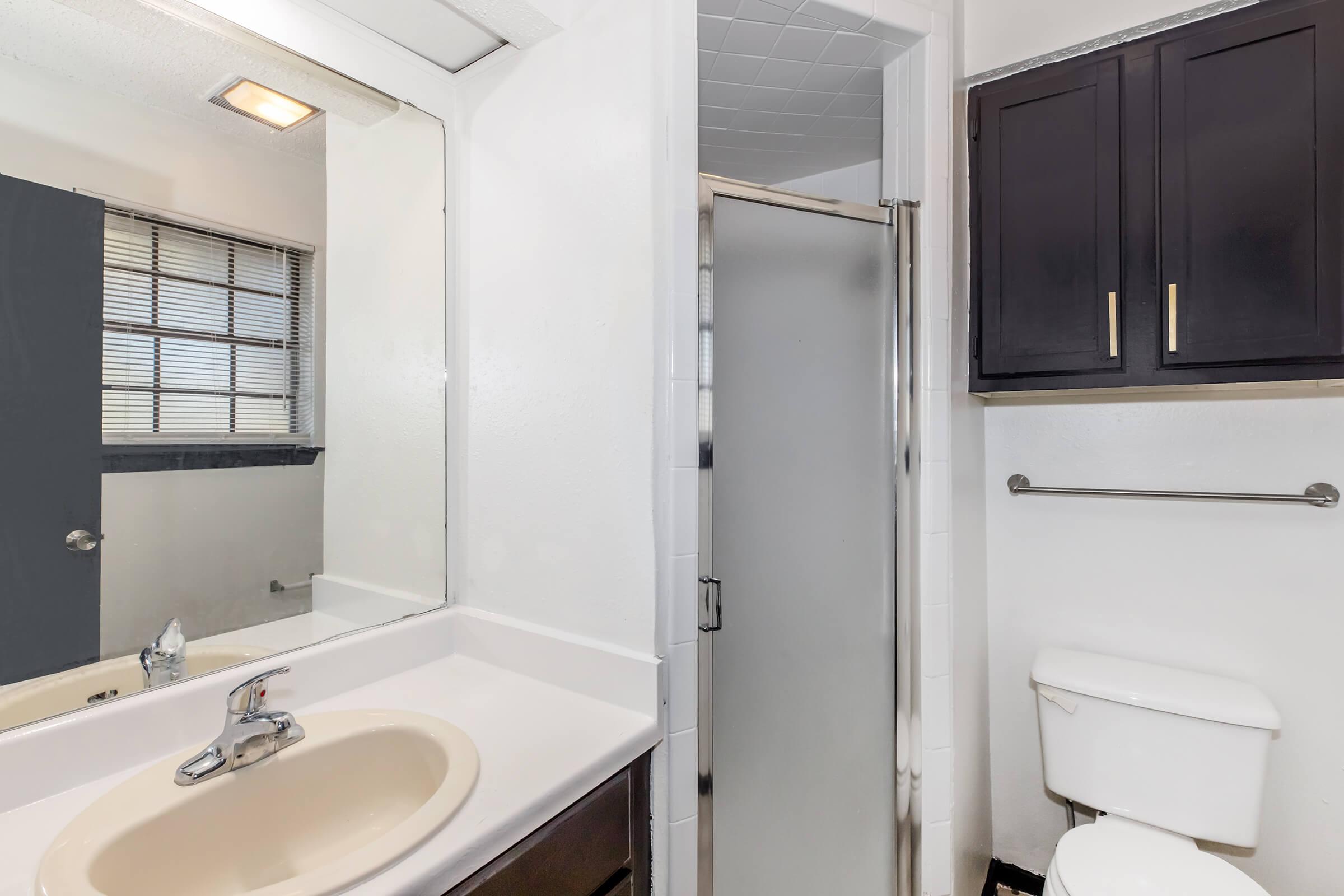 A bathroom featuring a wall-mounted mirror above a sink with a beige countertop, a shower stall with a glass door, a white toilet, and dark wood cabinetry. Natural light enters through a window with horizontal blinds. The walls are painted white, and the overall space is clean and well-maintained.