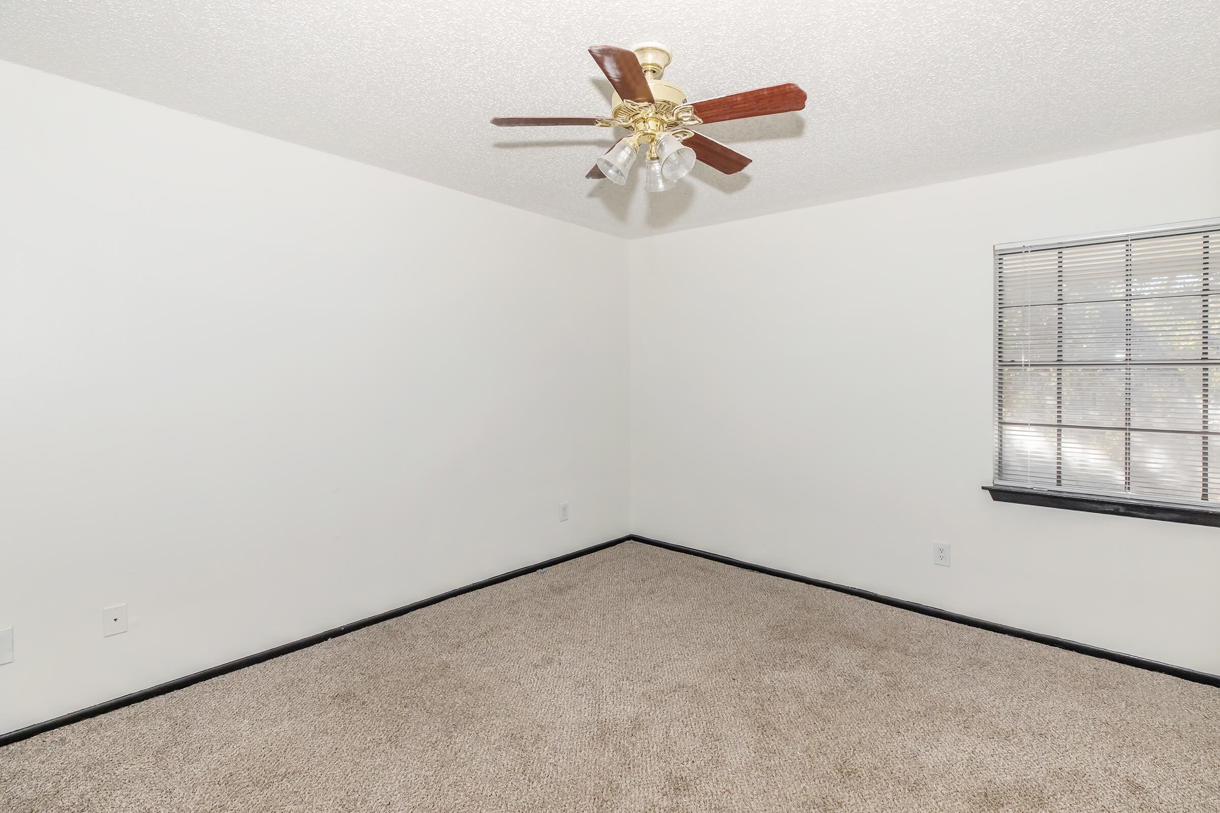 A light-colored room with beige carpet, a ceiling fan with wooden blades, and a window with multiple panes. The walls are plain and white, and the room appears empty and well-lit.