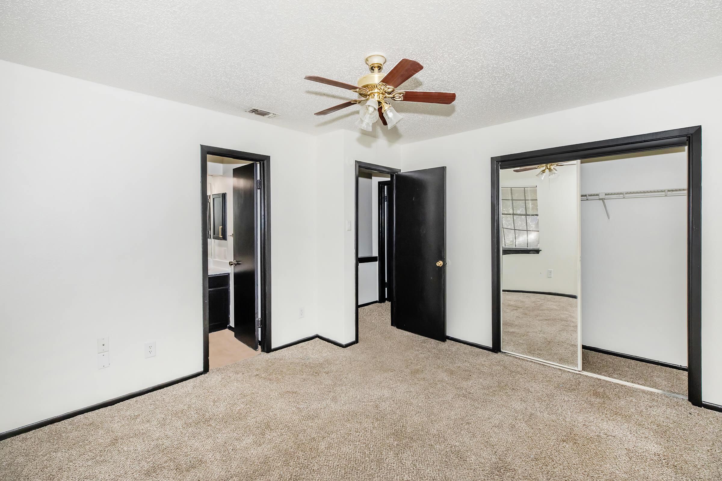 A spacious room featuring light beige carpet, neutral-colored walls, and a ceiling fan. Two doors lead to adjacent spaces, one opening to a bathroom area. A large mirror is mounted on one wall, reflecting the room's interior, which has natural light coming through a window.