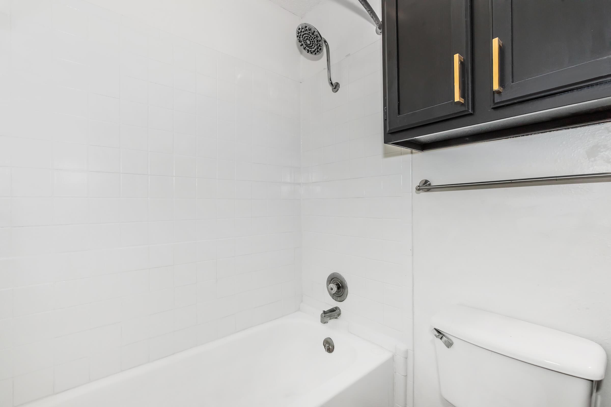 A clean and modern bathroom featuring a white bathtub and white tiled walls. The shower is mounted above the tub, and there is a sleek black cabinet with gold handles. A towel rack is visible, along with a white toilet on the side. The overall aesthetic is bright and minimalist.
