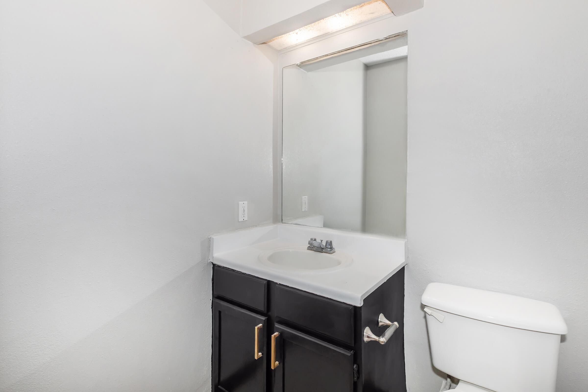 A small bathroom featuring a white sink with a black cabinet below, a light-colored countertop, a mirror above the sink, and a toilet beside it. The walls are painted in a light gray tone, creating a clean and minimalist look. Natural light may come from a window or light fixture above the mirror.