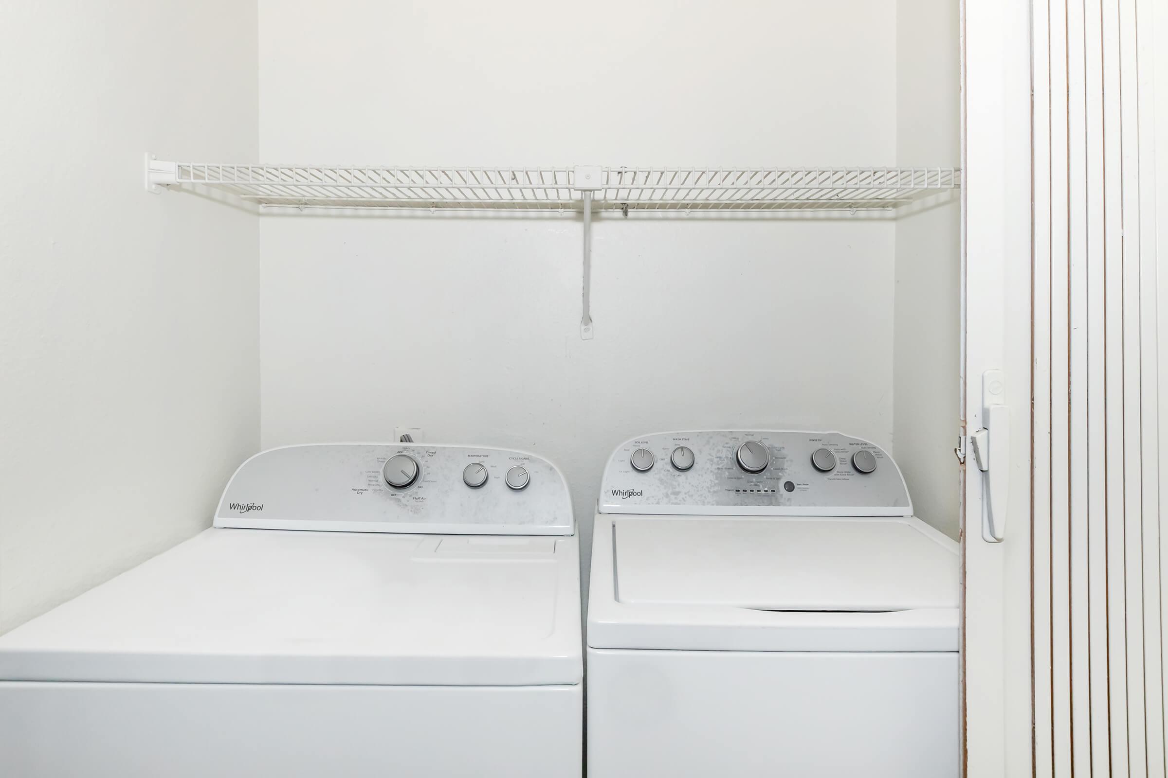 A clean laundry room featuring a white washing machine and dryer side by side, with control knobs and settings visible. Above them, there is a wire shelf mounted on the wall for storage. The walls are plain and light-colored, creating a minimalist look.