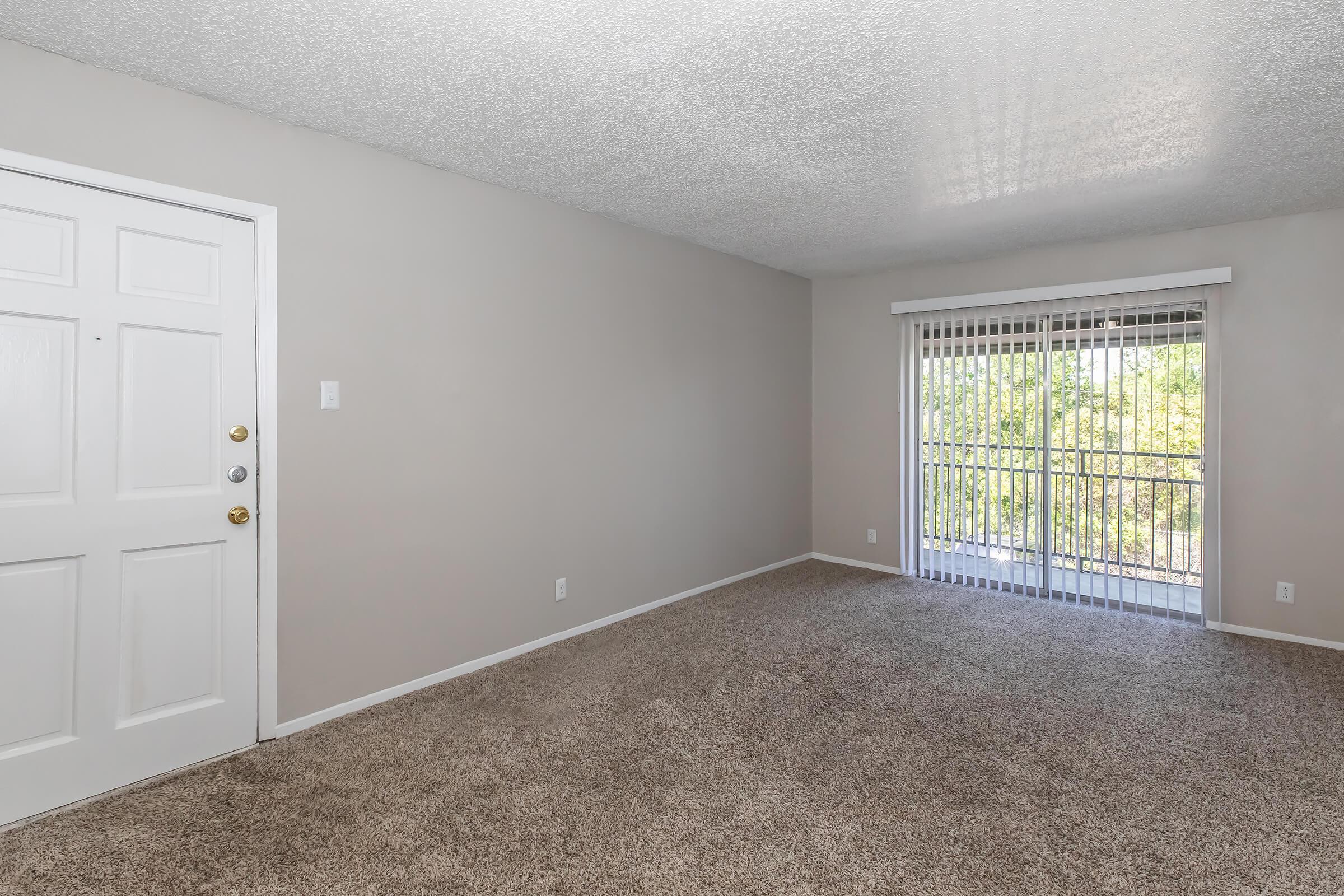 A spacious, empty room with light beige walls and carpet. There is a white door on the left and a sliding glass door on the right leading to a balcony. Natural light filters in, illuminating the area, which has a simple, uncluttered appearance.
