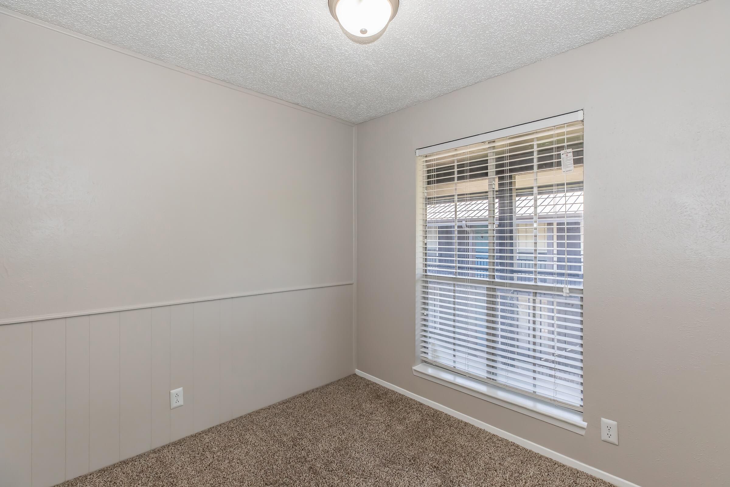 A well-lit interior room with beige walls and carpeted flooring. There is a window featuring white blinds that allows natural light. The ceiling has a light fixture, and the room appears to be freshly painted and tidy, suitable for various uses such as an office or bedroom.