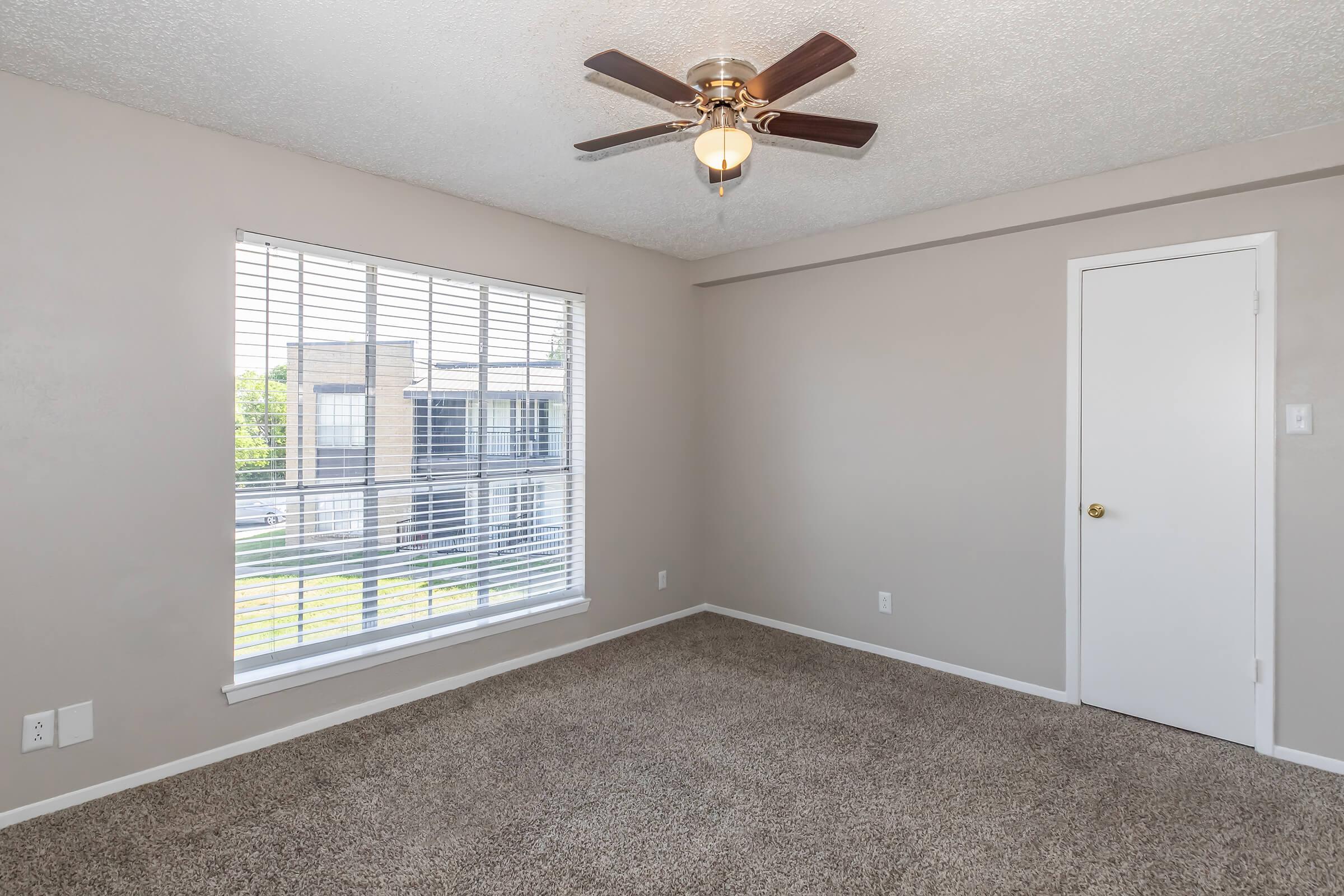 A well-lit, empty room with light beige walls and carpet flooring. A ceiling fan is located in the center, and a large window with white blinds allows natural light to filter in. A closed white door is visible, and outside the window, there are glimpses of greenery and nearby buildings.