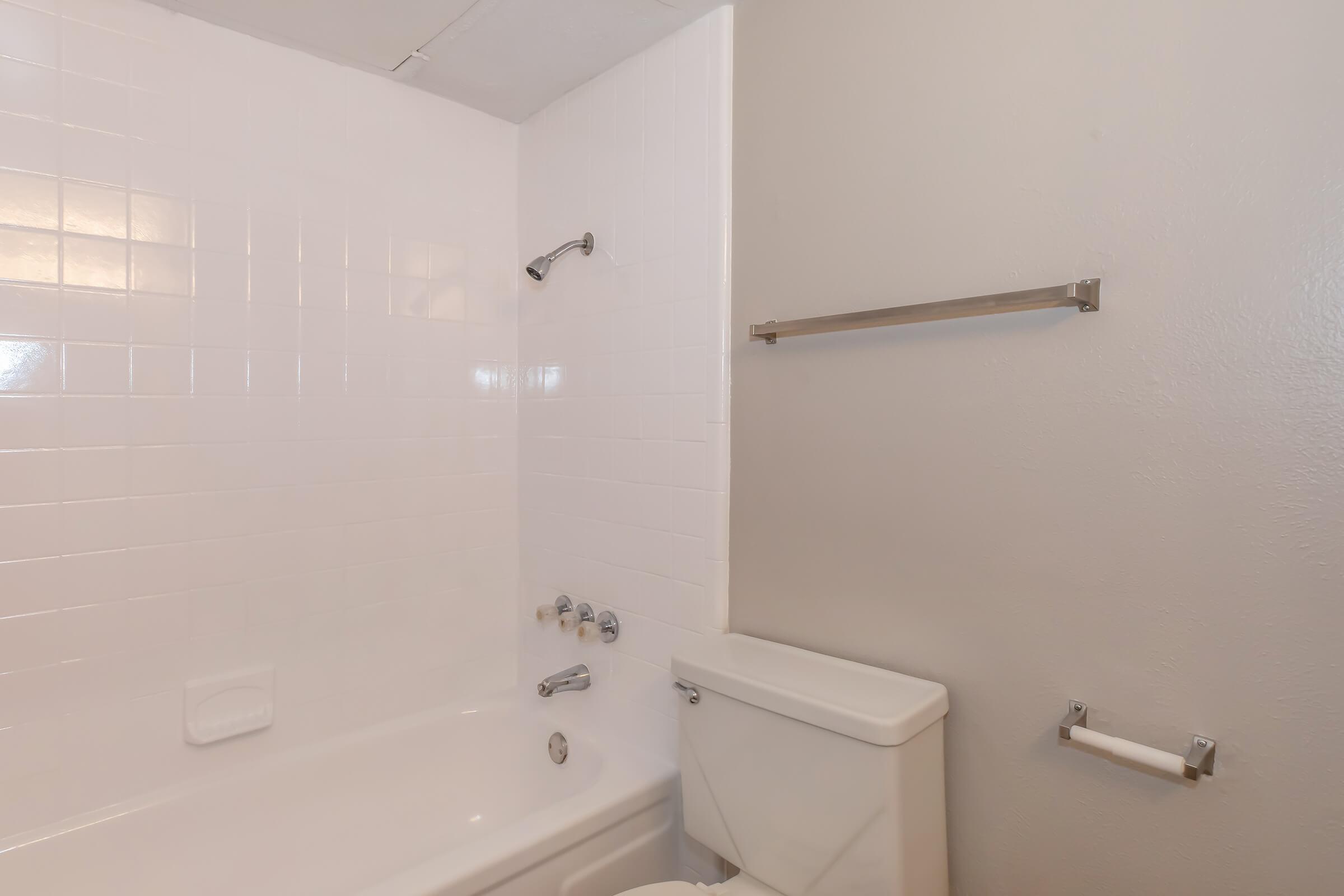 A clean, simple bathroom featuring a white tiled wall with a showerhead, a bathtub with faucet fixtures, a toilet, and a towel bar. The walls are painted a light color, and the overall space looks well-maintained and functional.