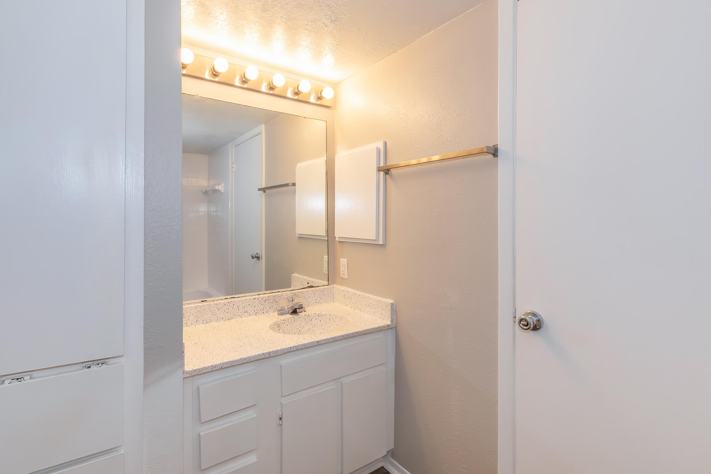 A well-lit bathroom featuring a white vanity with a granite countertop, a mirror above it, and a towel rack on the wall. The walls are painted in a light gray color. A door leads to another area, and the overall space appears clean and modern.