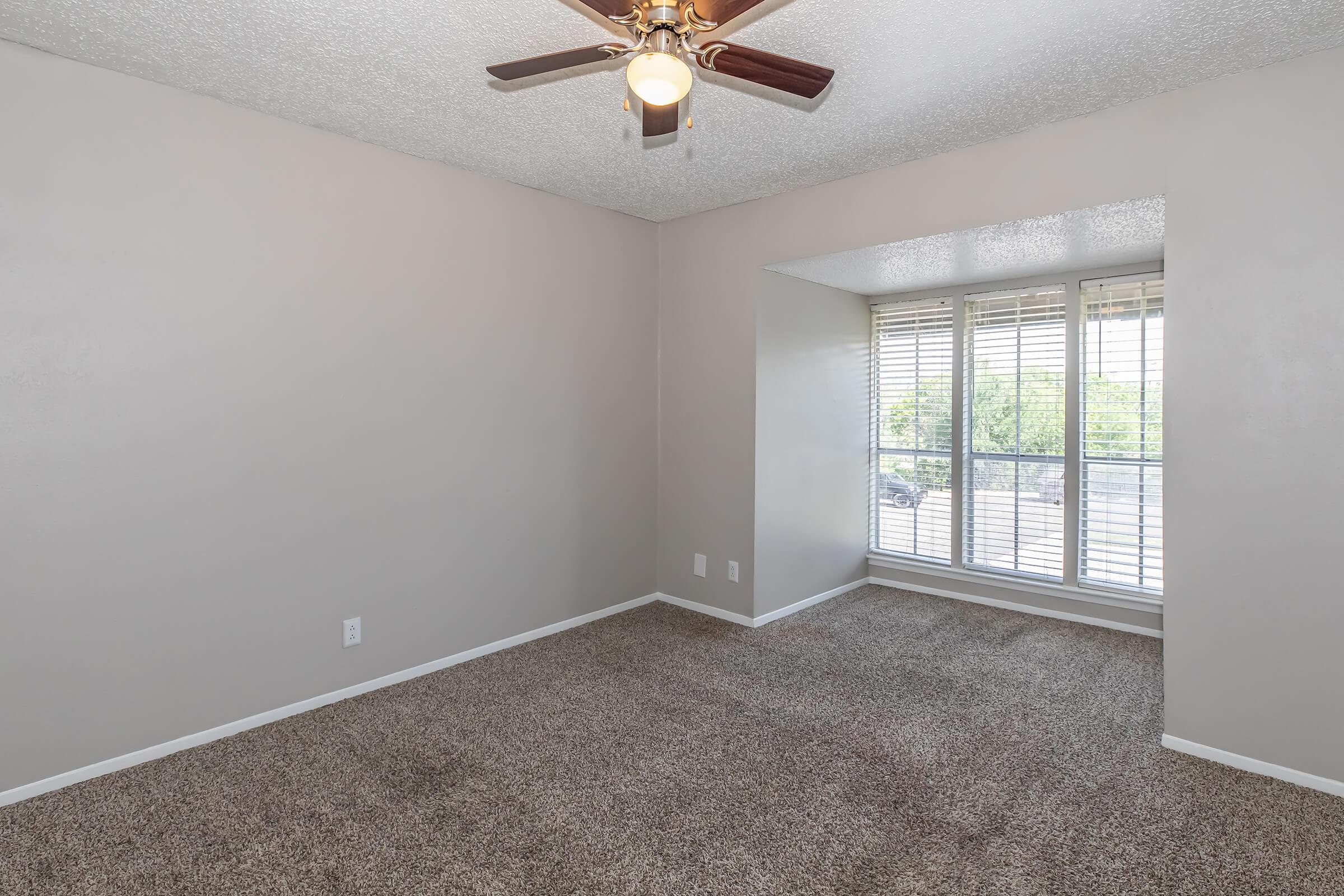 A spacious living area with light beige walls and carpet. A ceiling fan is centered in the room, and there is a large window with blinds allowing natural light. The space appears clean and inviting, suitable for various furniture arrangements.