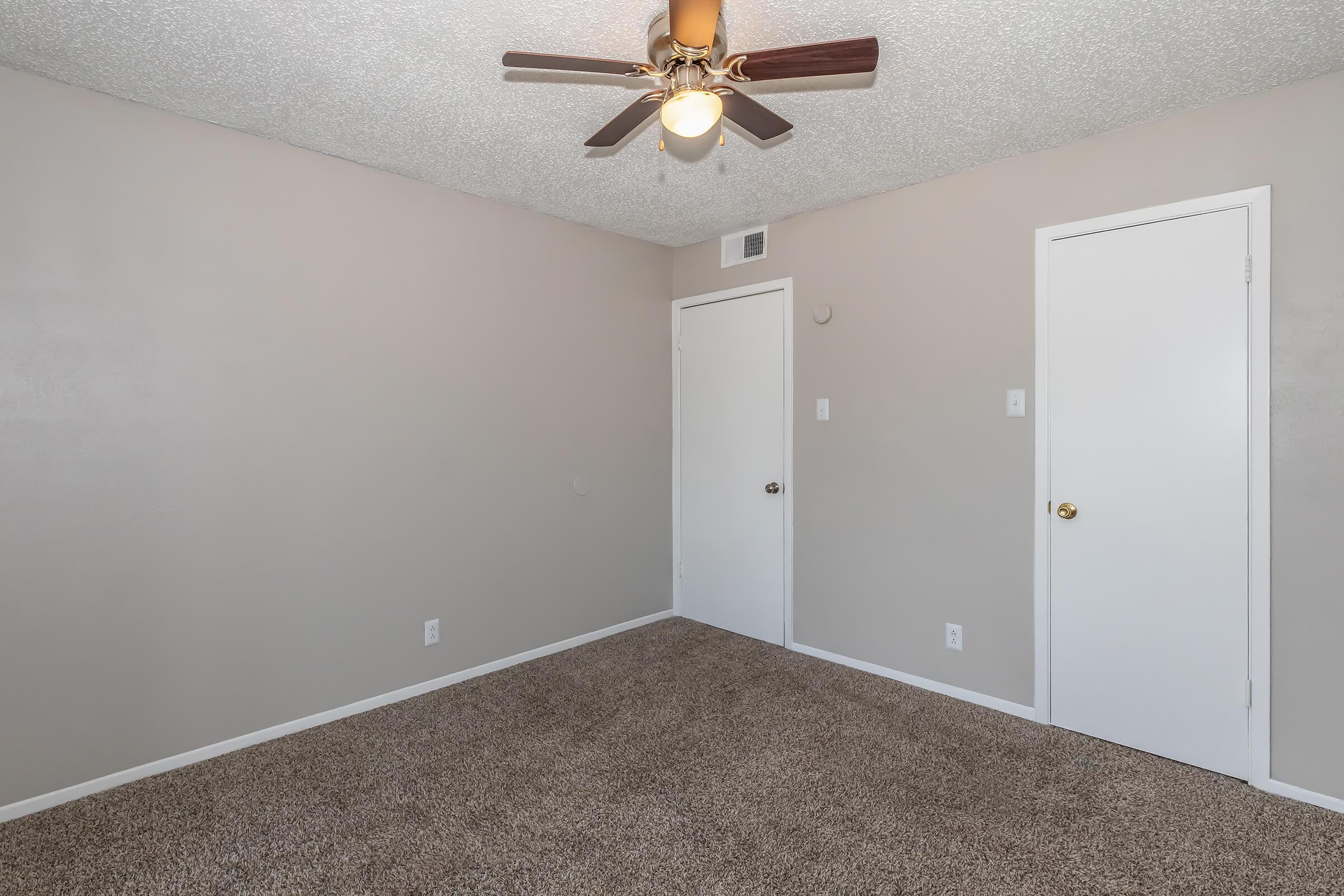 A simple, empty room with light beige walls and a textured ceiling. The floor is covered in brown carpeting. A ceiling fan with light is mounted in the center. There are two closed doors on either side, one with a round doorknob. The room is well-lit and neutral in color, creating a spacious feel.