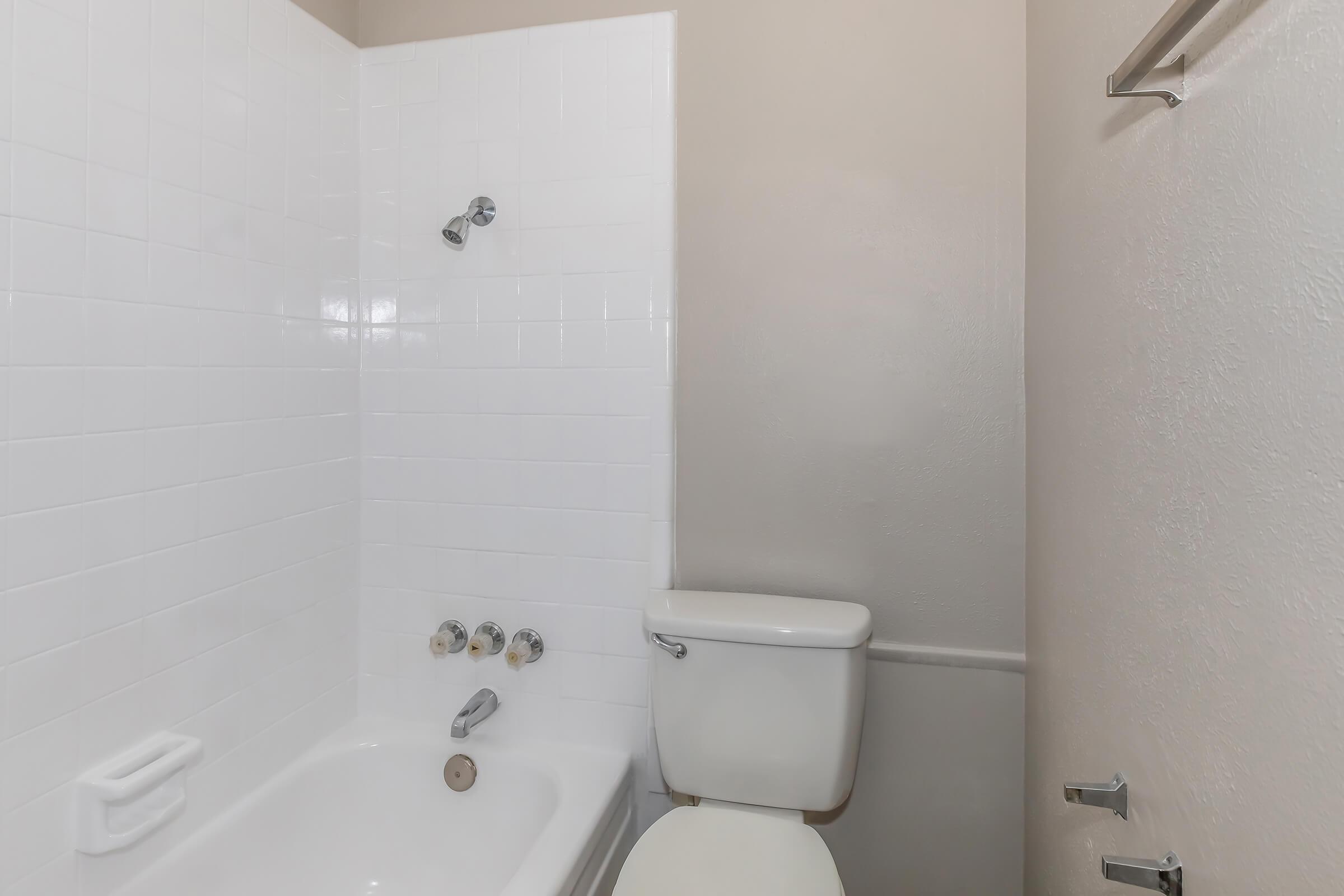 A clean, minimalistic bathroom featuring a white bathtub with a showerhead above, next to a white toilet. The walls are light-colored, and the tiles are white, creating a bright and spacious feel. There are simple fixtures and a towel rack mounted on the wall.