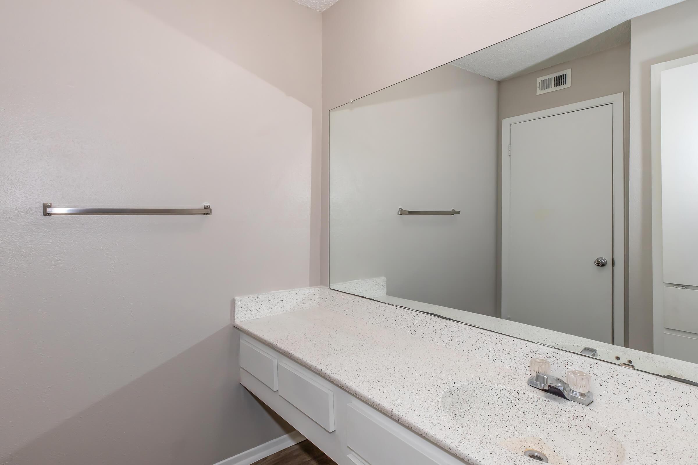 A clean and modern bathroom with a light-colored wall, a large mirror above a speckled countertop, and a sink. The countertop features two sink fixtures, and there are two towel bars mounted on the wall. A white door is visible in the background, adding to the simple and functional design.