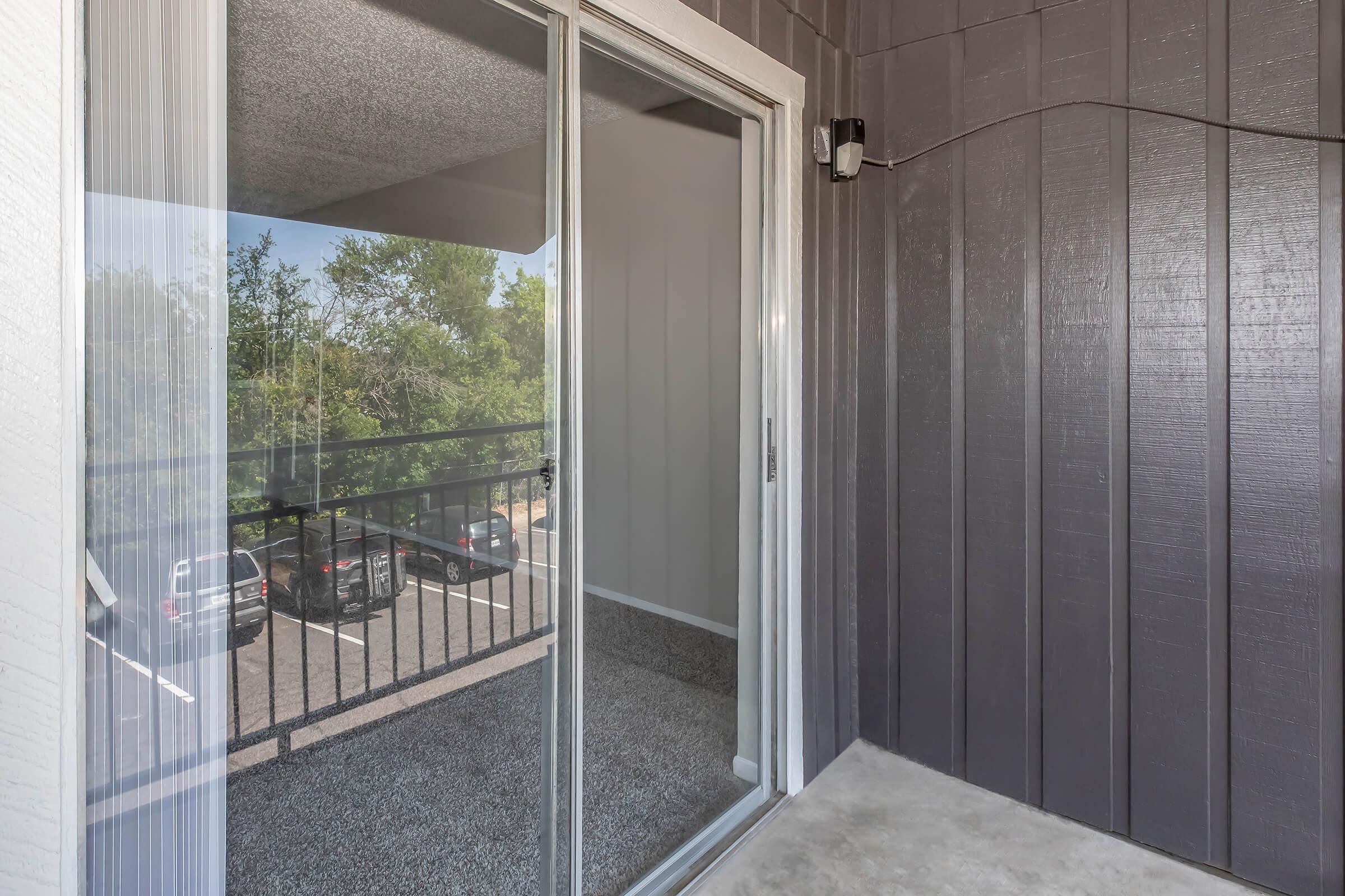 A view from inside an apartment balcony door, showing a sliding glass door leading to an outdoor space. The balcony features a railing with a view of parked cars below and greenery outside. The interior walls are gray, and the floor is carpeted.