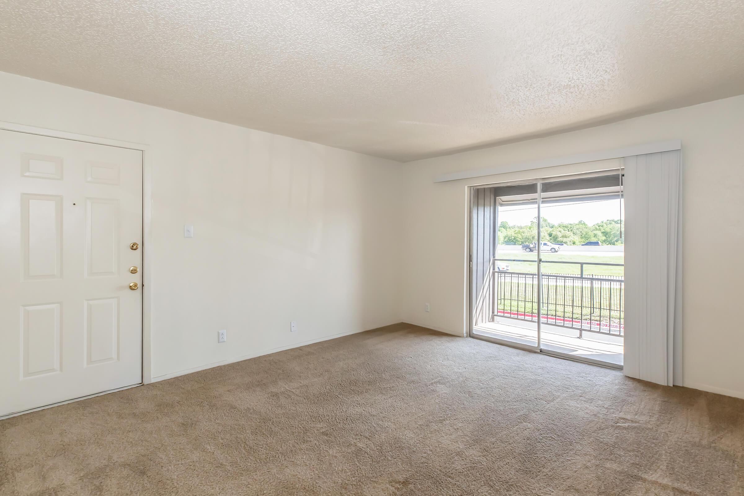 A spacious, empty room with beige carpet and white walls. A door on the left leads outside, and a large sliding glass door on the right opens to a balcony with a view of greenery. Natural light floods the room, creating a bright and airy atmosphere.