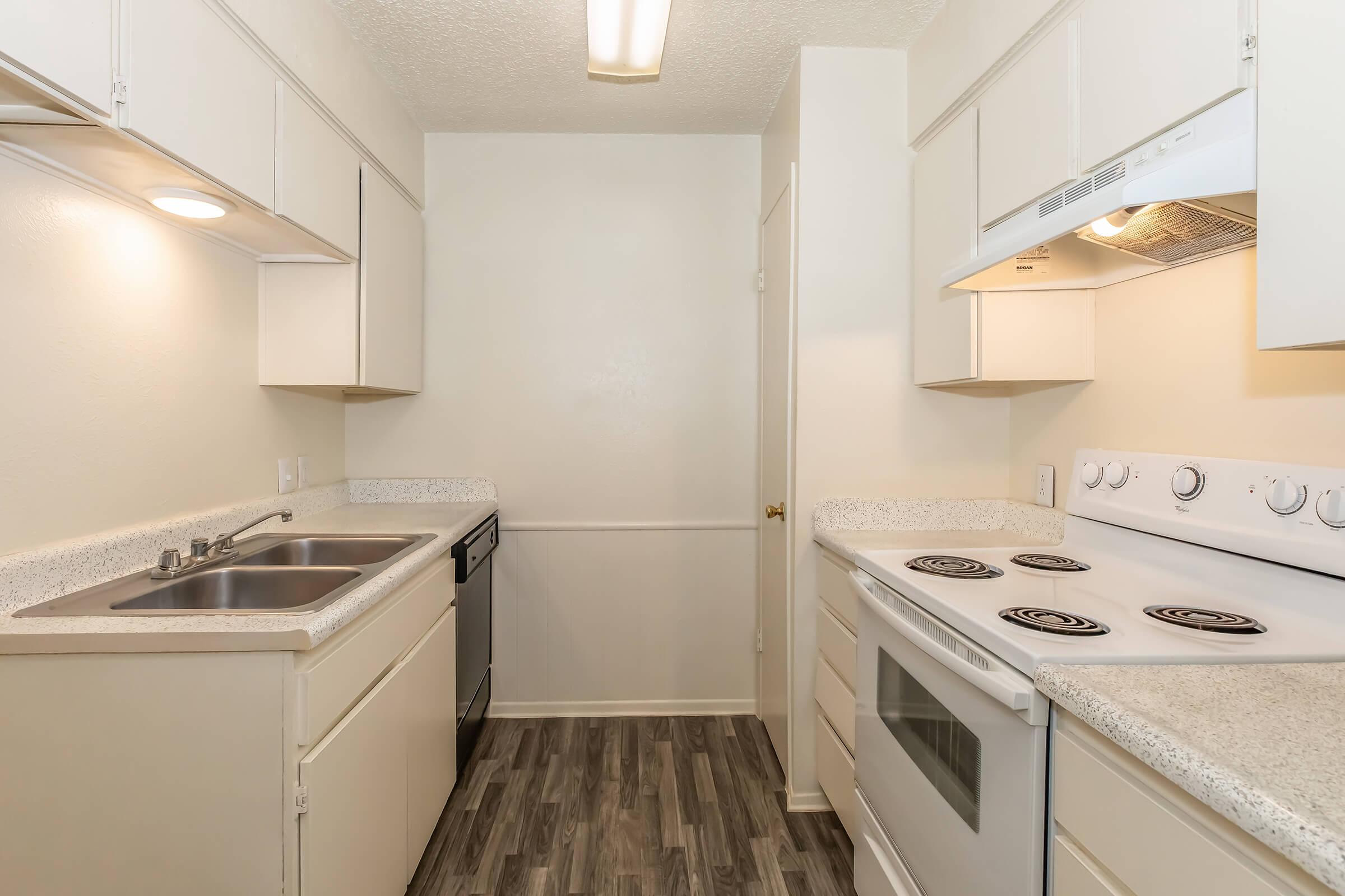 A small, modern kitchen featuring white cabinetry, a double sink, a dishwasher, a stovetop, and an oven. The countertops are light-colored with a speckled design, and the flooring is a wood-like laminate. The walls are painted a neutral color, enhancing the bright and clean appearance of the space.
