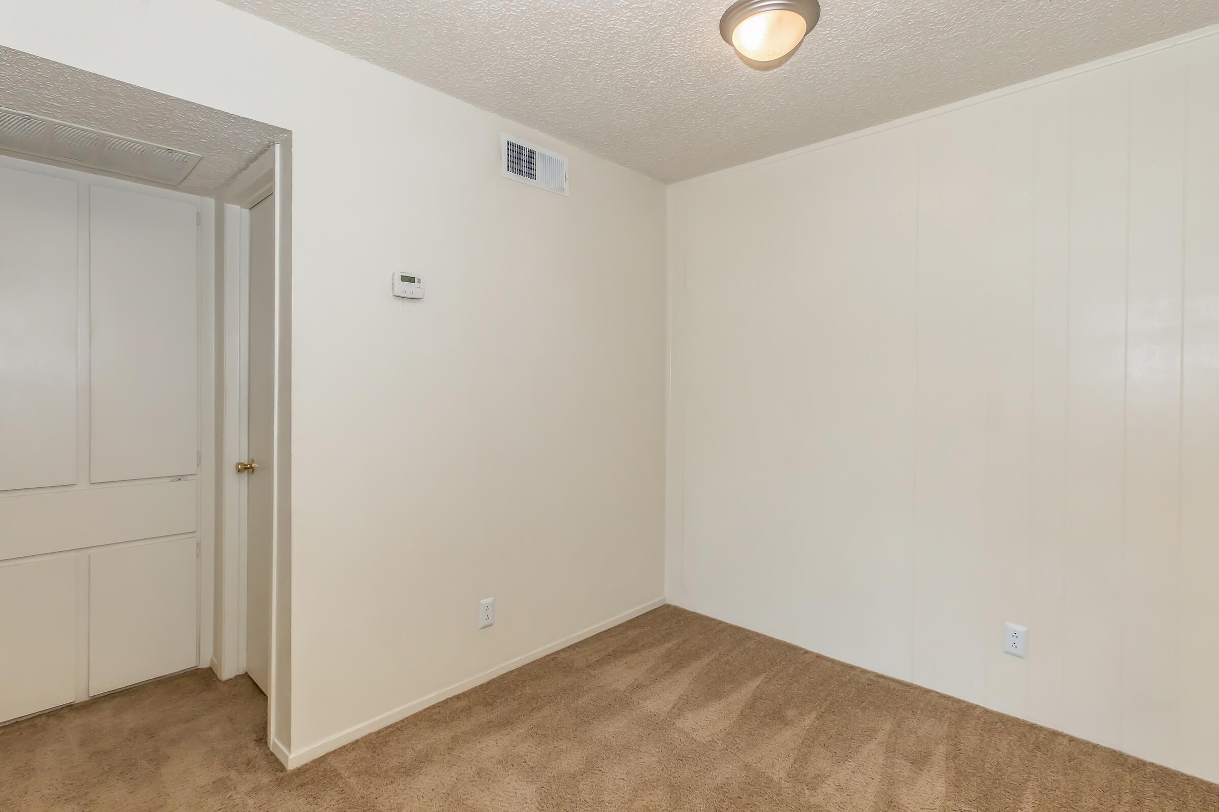 Empty room with light beige carpet and white walls. A door on the left leads to another space, and a small wall-mounted thermostat is visible. A ceiling light fixture provides illumination. The overall atmosphere is neutral and clean, suitable for various uses.