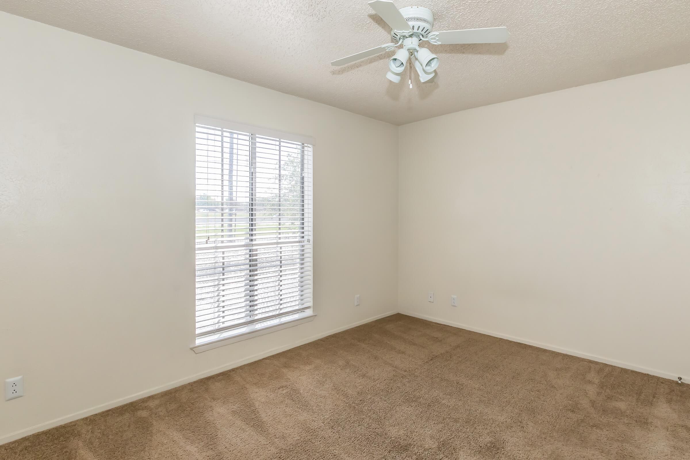 Empty room with beige carpet, light-colored walls, and a ceiling fan. A window with white blinds allows natural light to enter the space. The room appears clean and well-maintained, suitable for various uses, such as a bedroom or office.