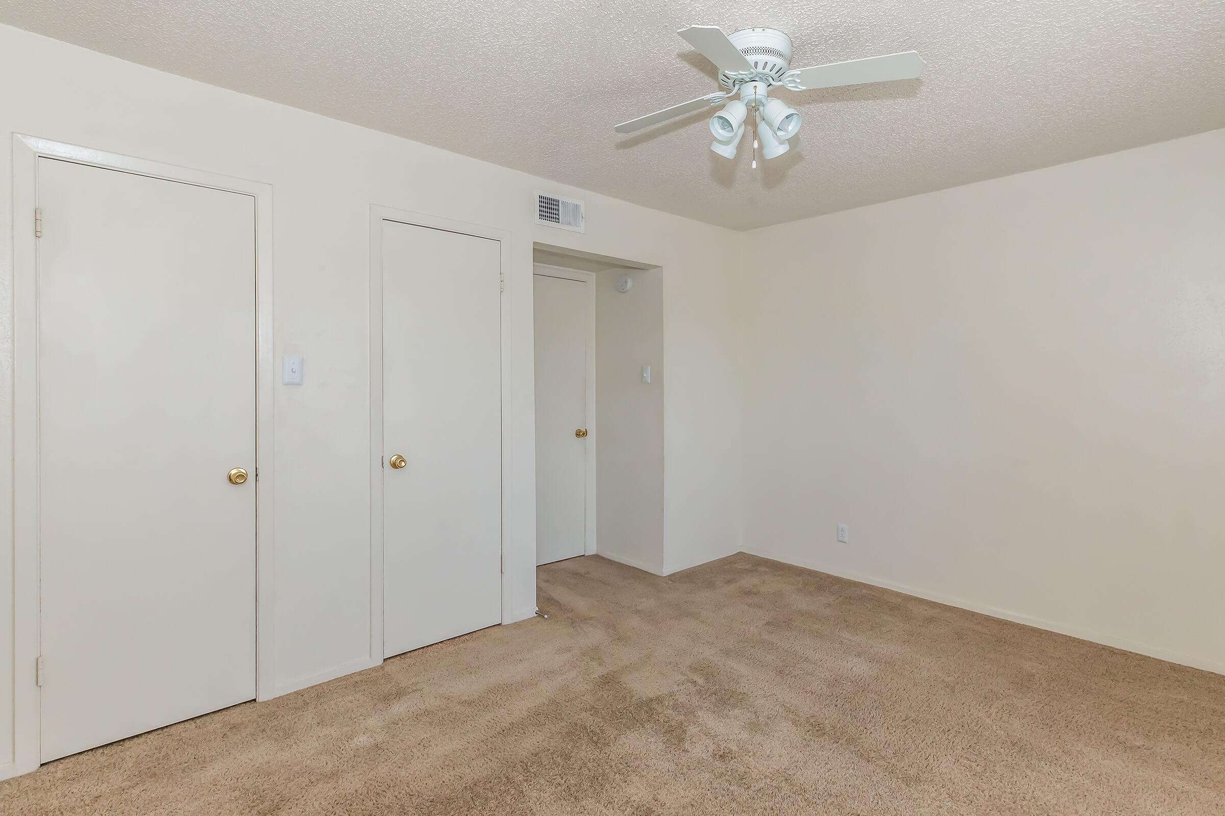 A spacious and empty room featuring light beige walls and carpeting. Two closed white doors are on the left side, and an additional door is visible in the rear. A ceiling fan with white blades hangs from the ceiling, providing ventilation. The overall ambiance is neutral and inviting.