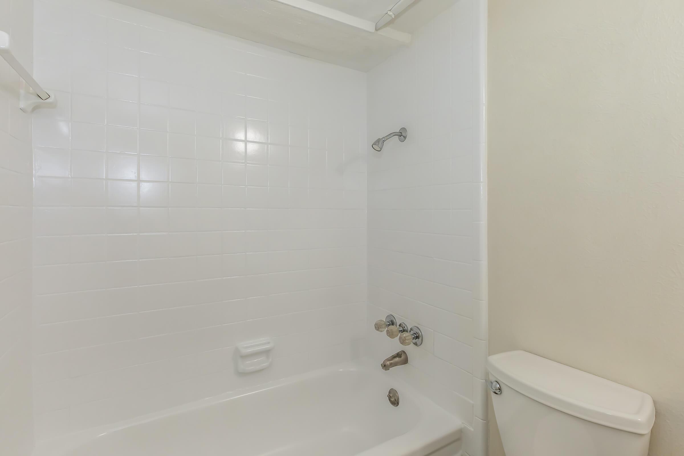 A clean and simple bathroom featuring a white tiled shower area with a bathtub, a showerhead, and control knobs. A toilet is positioned adjacent to the bathtub, also in a white finish. The overall aesthetic is bright and minimalistic, with neutral-toned walls.