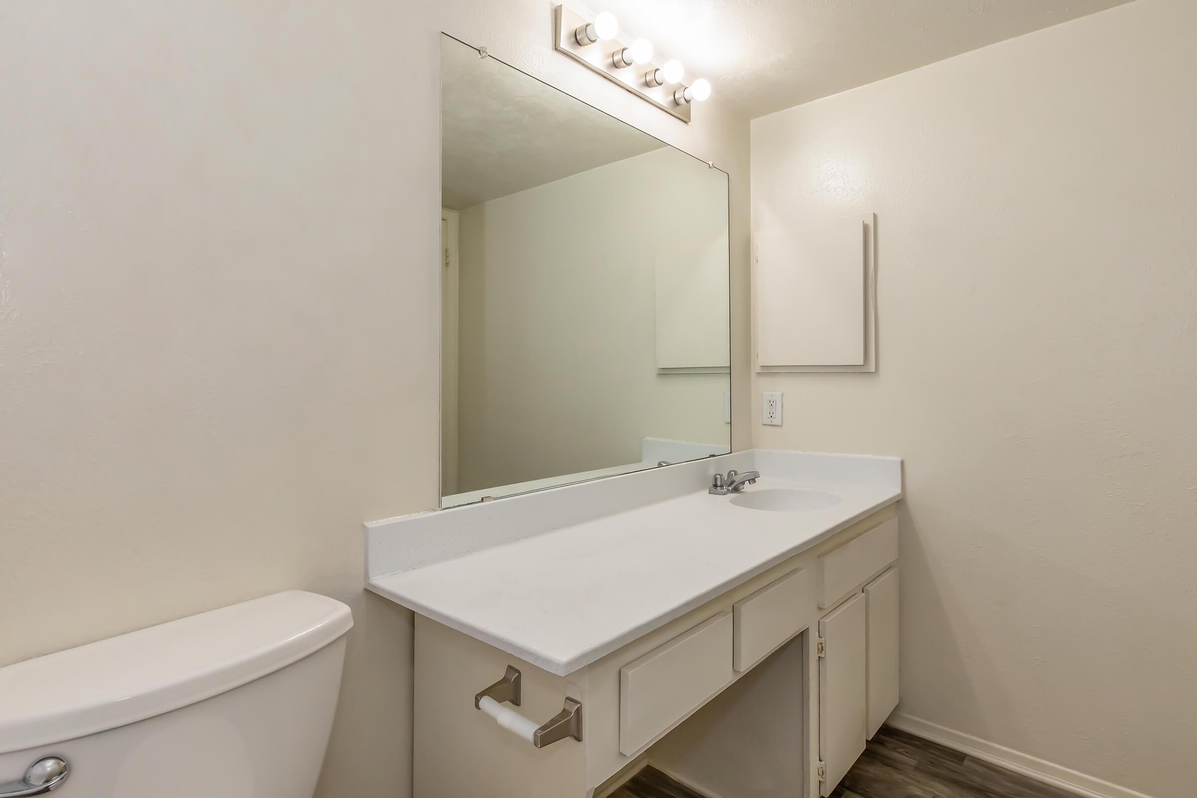 A clean, modern bathroom featuring a white countertop with a sink, a large mirror above, and stylish lighting. A toilet is positioned on the left side. The walls are painted in a light color, and the flooring is a dark wood style, creating a bright and inviting atmosphere.