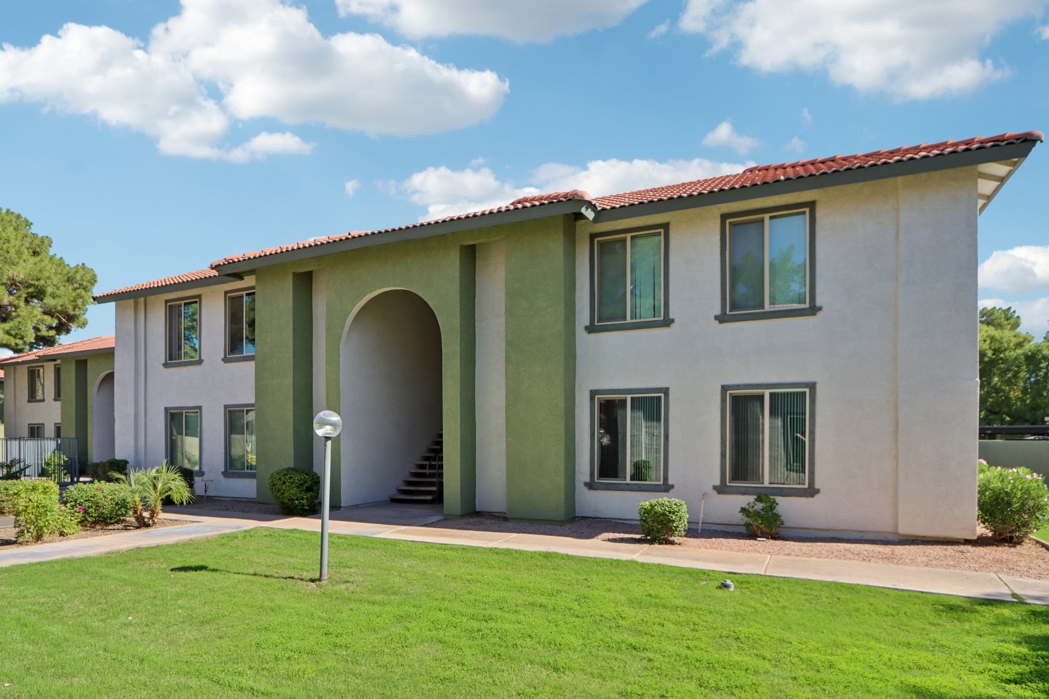 Two-story apartment building with a green and white facade. The entrance features an archway, and there are large windows on either side. Well-maintained landscaping includes shrubs and grass in the foreground. A circular light fixture stands along the pathway leading to the entrance, under a partly cloudy sky.