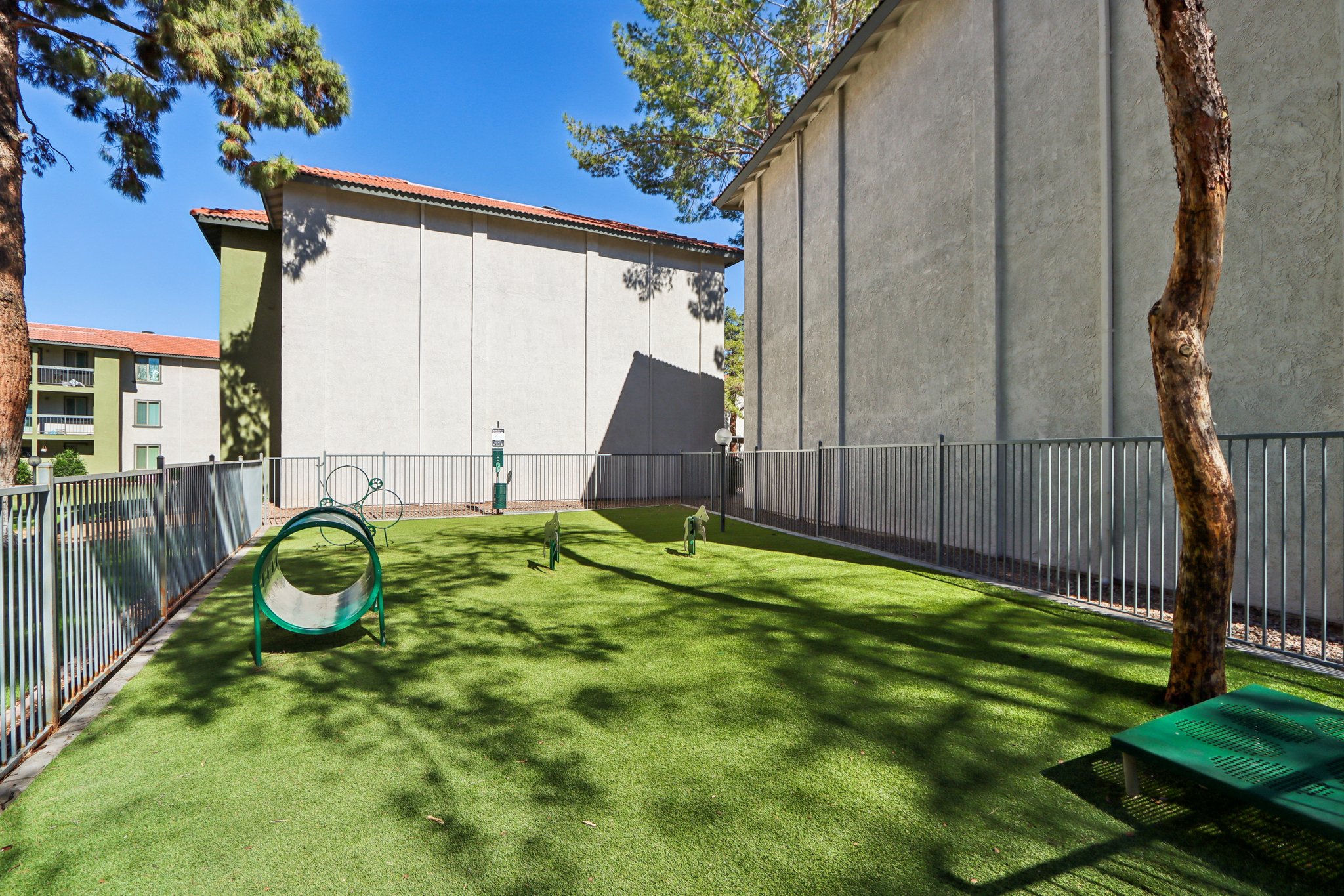 A green outdoor space featuring artificial turf, surrounded by tall trees and buildings. There is a tunnel structure, small agility equipment, and a shaded area, making it suitable for pets, particularly dogs, to play and exercise. The clear blue sky overhead enhances the bright atmosphere.