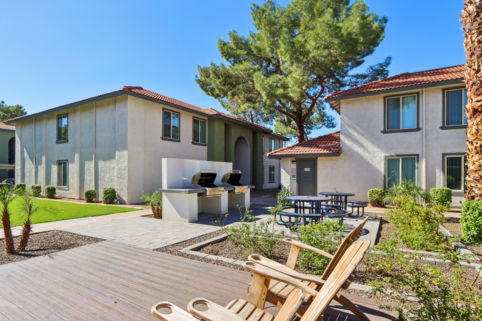 Exterior view of a residential complex featuring two buildings with a stucco finish and red tile roofs. In the foreground, there are wooden lounge chairs and a landscaped area with palm trees. A barbecue station is visible, along with picnic tables, creating a communal outdoor space for residents.