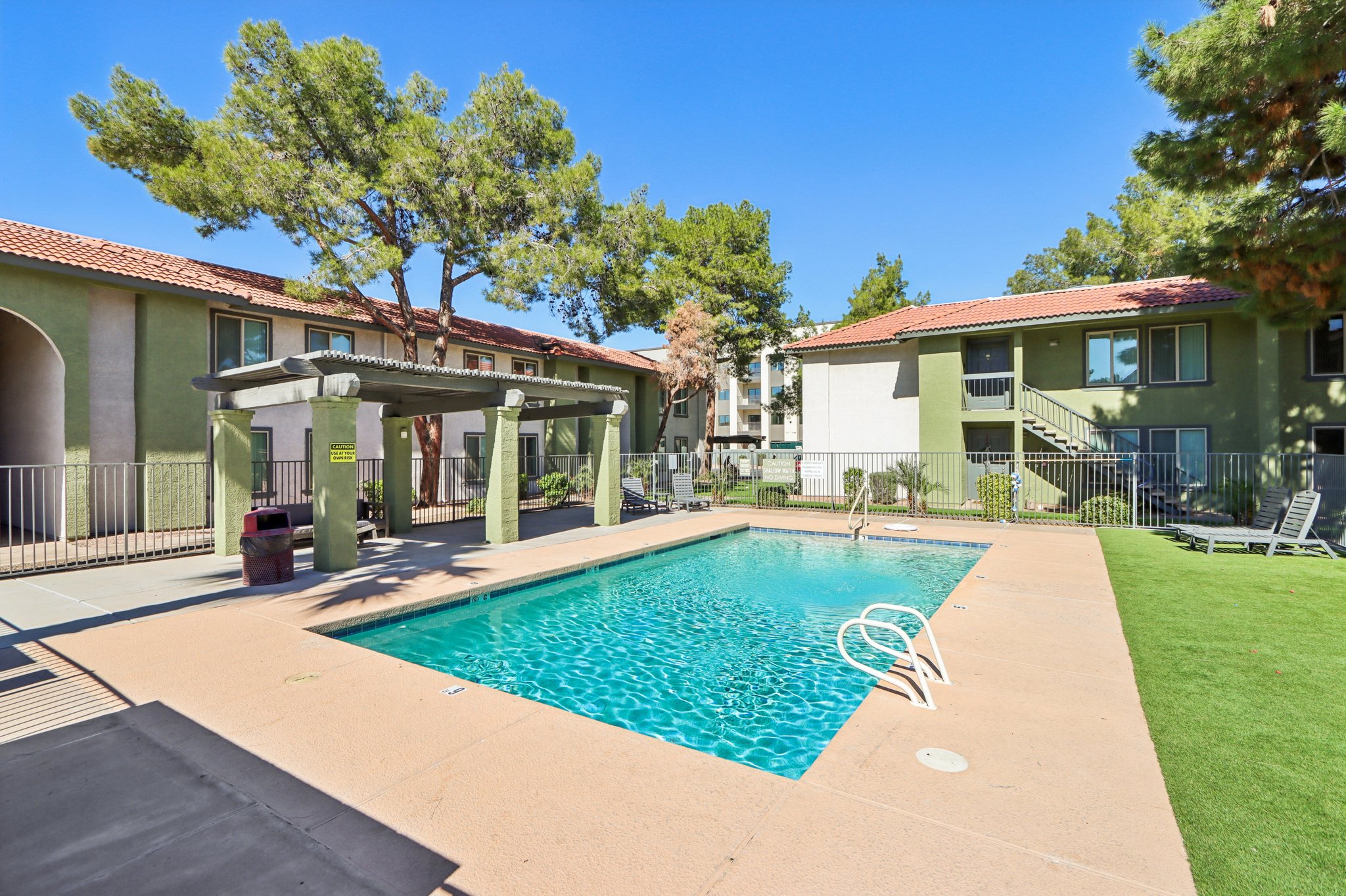 A clear swimming pool surrounded by a patio area with lounge chairs. The pool is framed by green grass and tall trees. In the background, there are two residential buildings with balconies and a sunny blue sky.