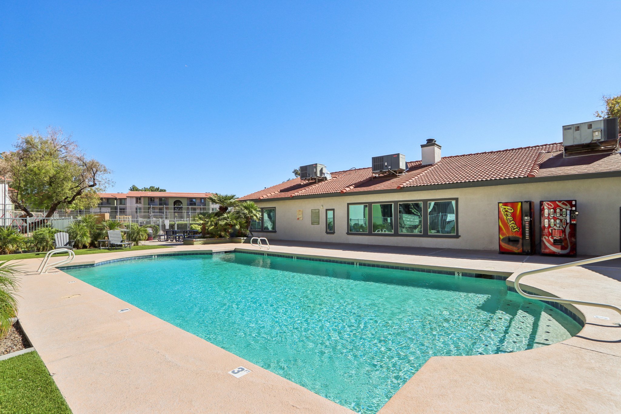 A clear swimming pool surrounded by a patio area with lounge chairs and greenery. In the background, there are two buildings, one with large windows and a vending machine. The sky is bright blue, and the setting appears sunny and inviting.