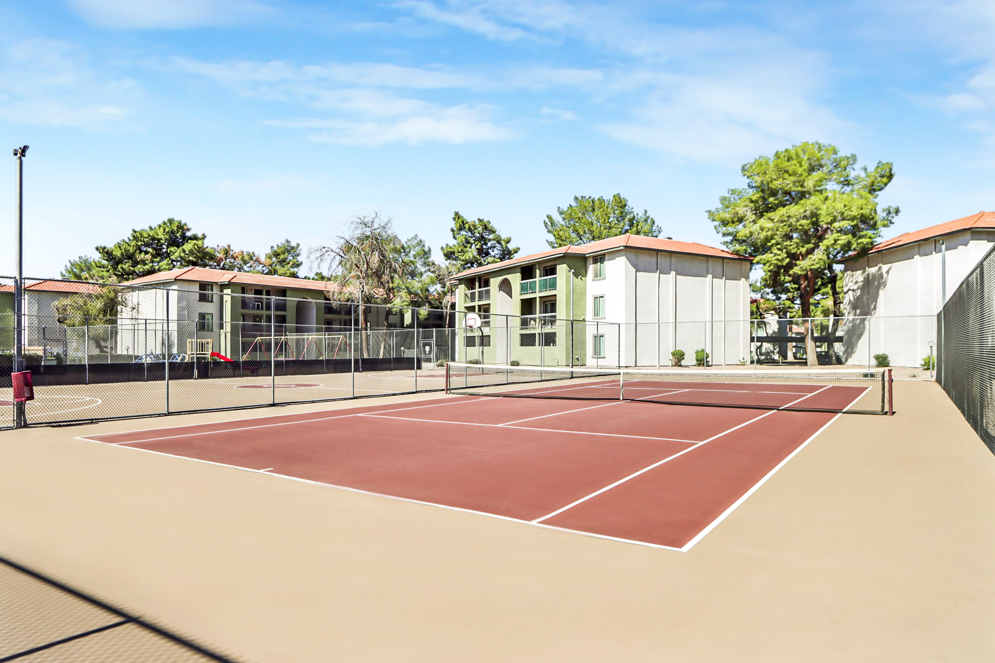 Tennis courts with a reddish-brown surface surrounded by a black fence. In the background, there are residential buildings with green trees, under a clear blue sky. The scene captures a bright, sunny day, providing a serene atmosphere for sports and recreation.