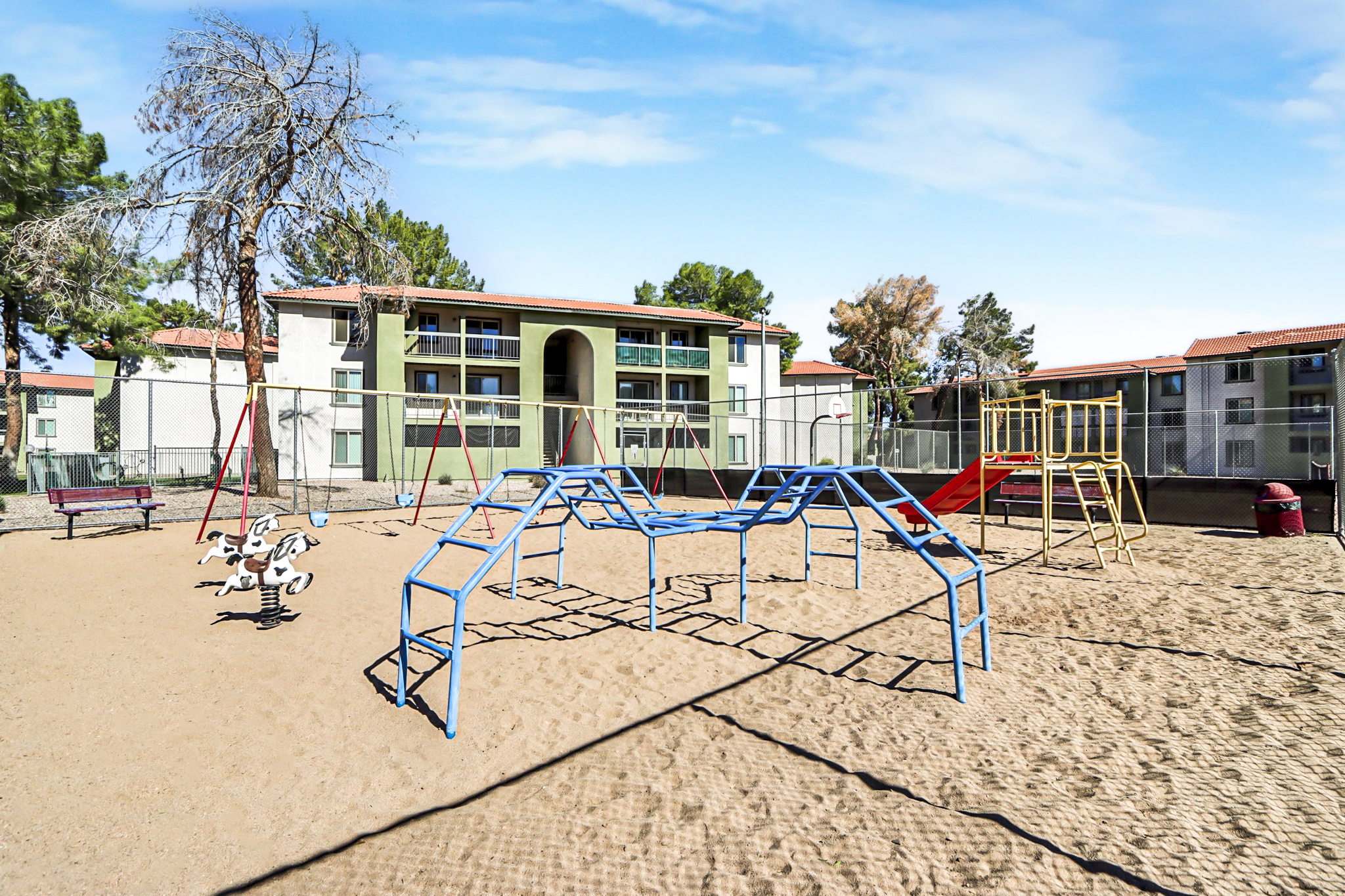A sandy playground featuring various colorful equipment, including swings, a climbing structure, and a monkey bar set, surrounded by a fence. In the background, there are multi-story residential buildings with green and beige exteriors and trees. The sky is clear and blue.