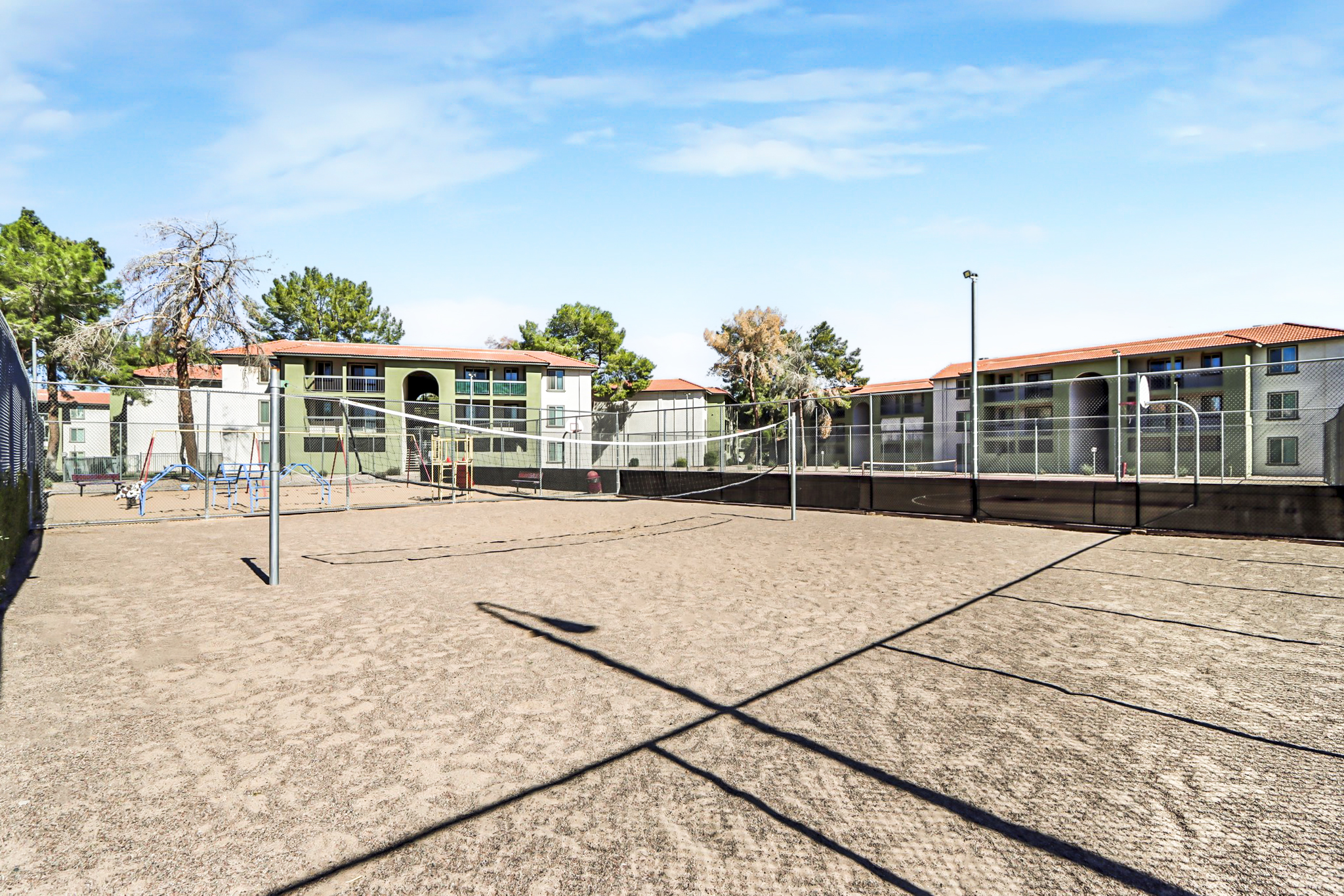 A sandy volleyball court surrounded by a fence, with two volleyball nets. In the background, there are residential buildings and trees under a clear blue sky. The scene is sunny and inviting, ideal for outdoor sports activities.