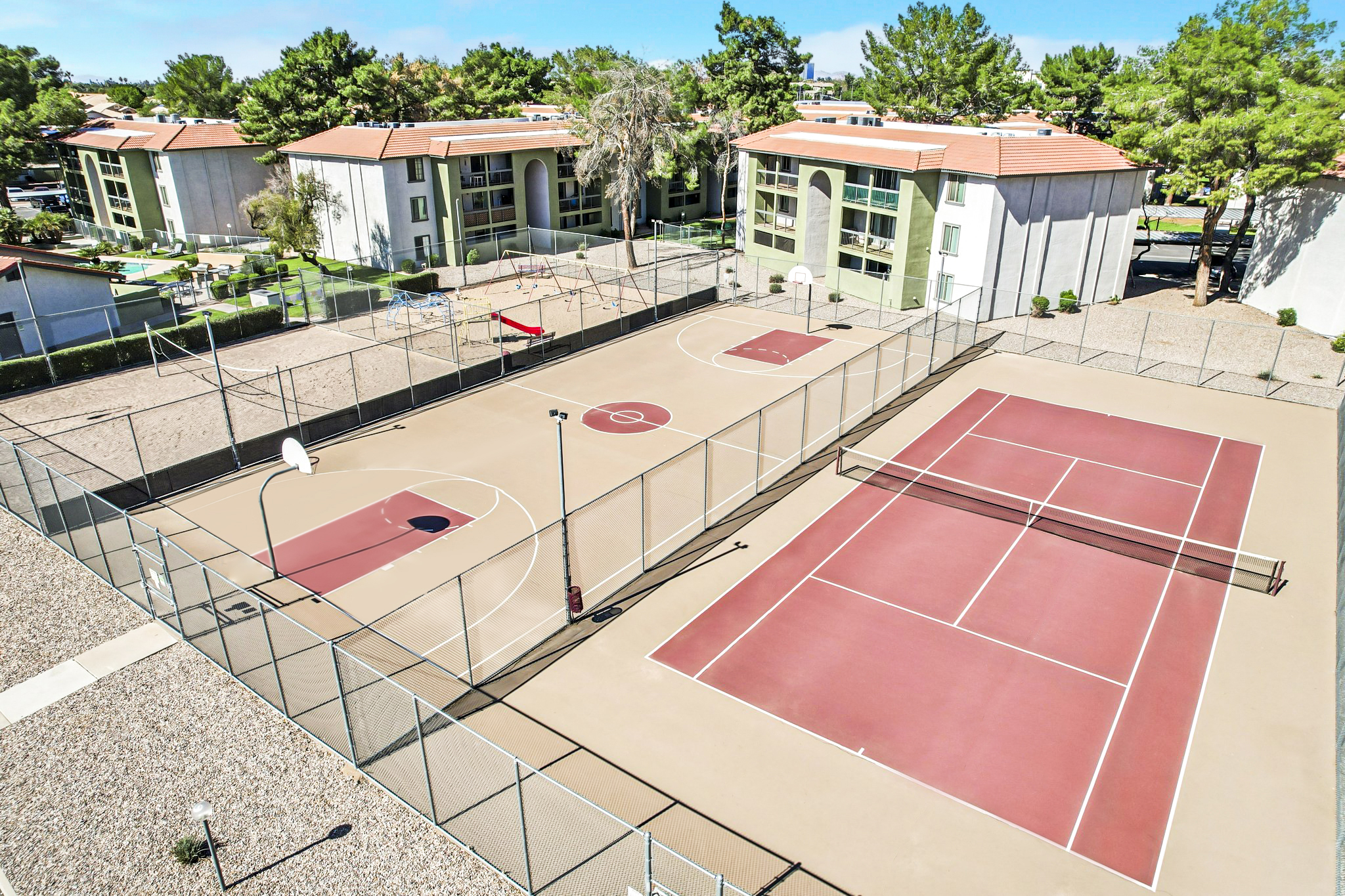Aerial view of a sports complex featuring two tennis courts and a basketball court. The courts are surrounded by a fence and bordered by residential buildings with greenery. The surface of the courts is predominantly red, with basketball hoops at each end and a net dividing the tennis courts.