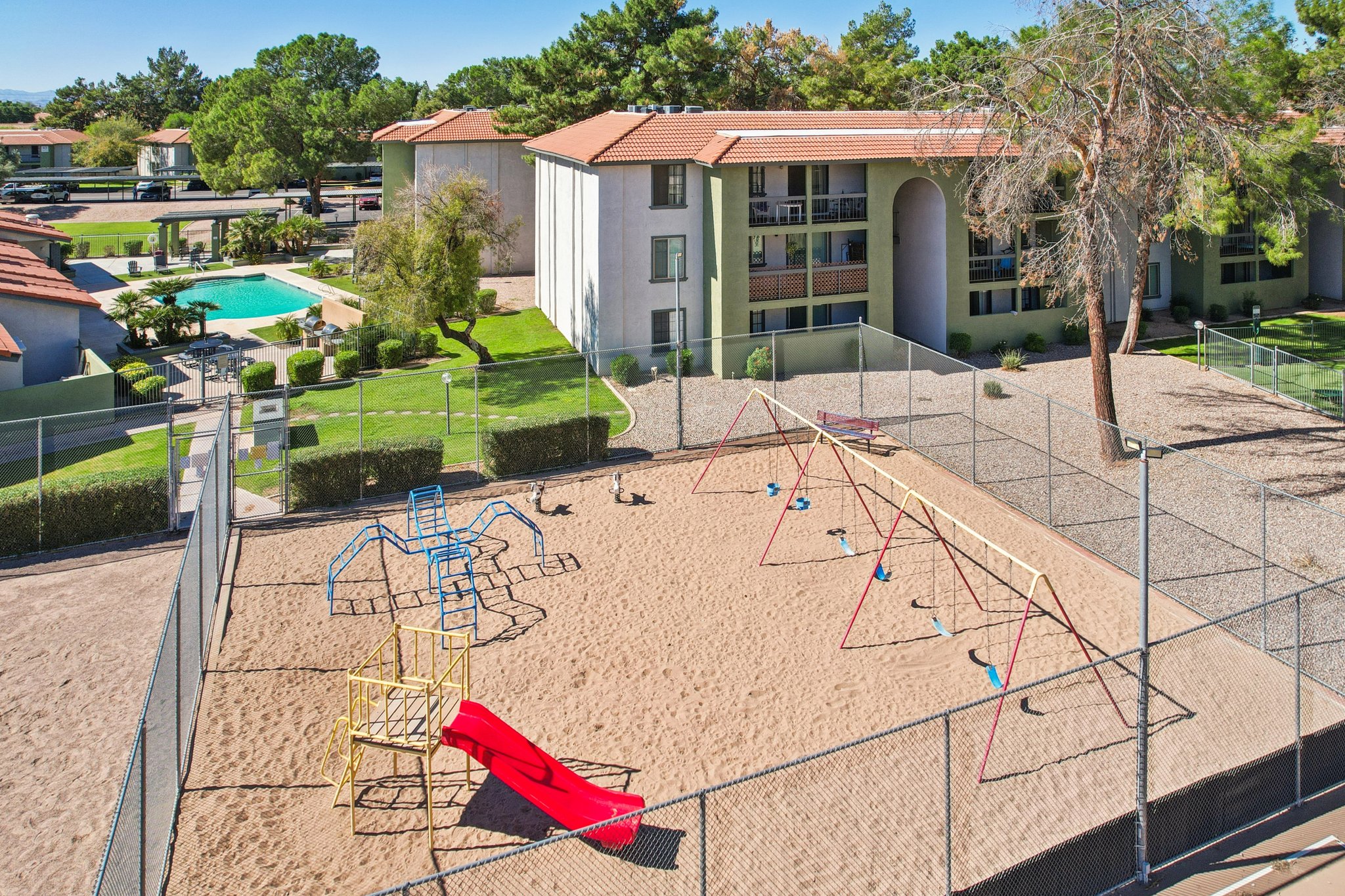 A sunny playground area enclosed by a fence, featuring a yellow climbing structure and a red slide. Nearby, there are several swings on a sandy surface. In the background, there are residential buildings and a swimming pool surrounded by greenery.