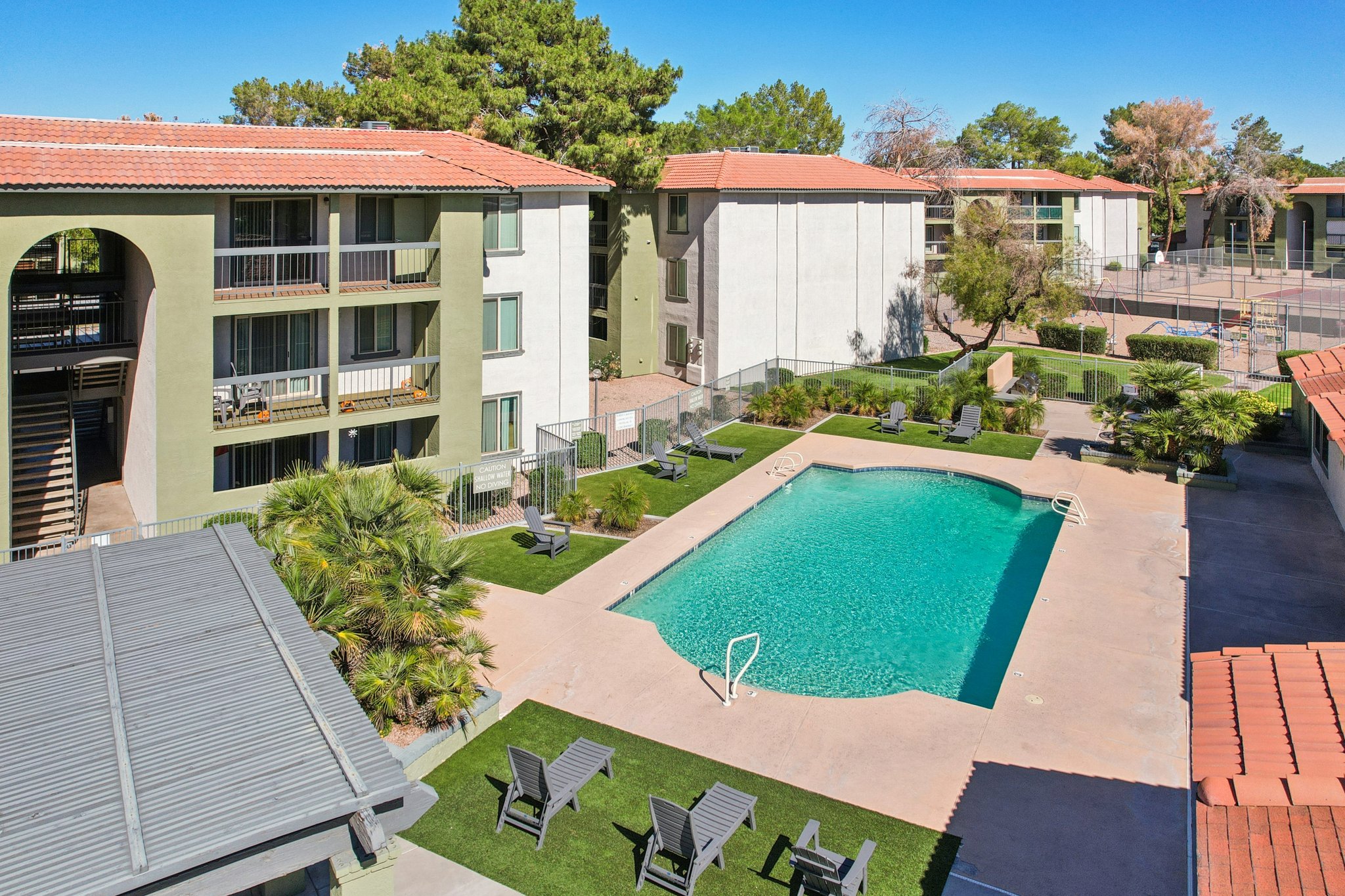 An aerial view of an apartment complex featuring several buildings with terracotta roofs. In the center, there is a sparkling blue swimming pool surrounded by lush landscaping, lounge chairs, and palm trees. The scene is set on a clear day with a bright blue sky and green trees in the background.