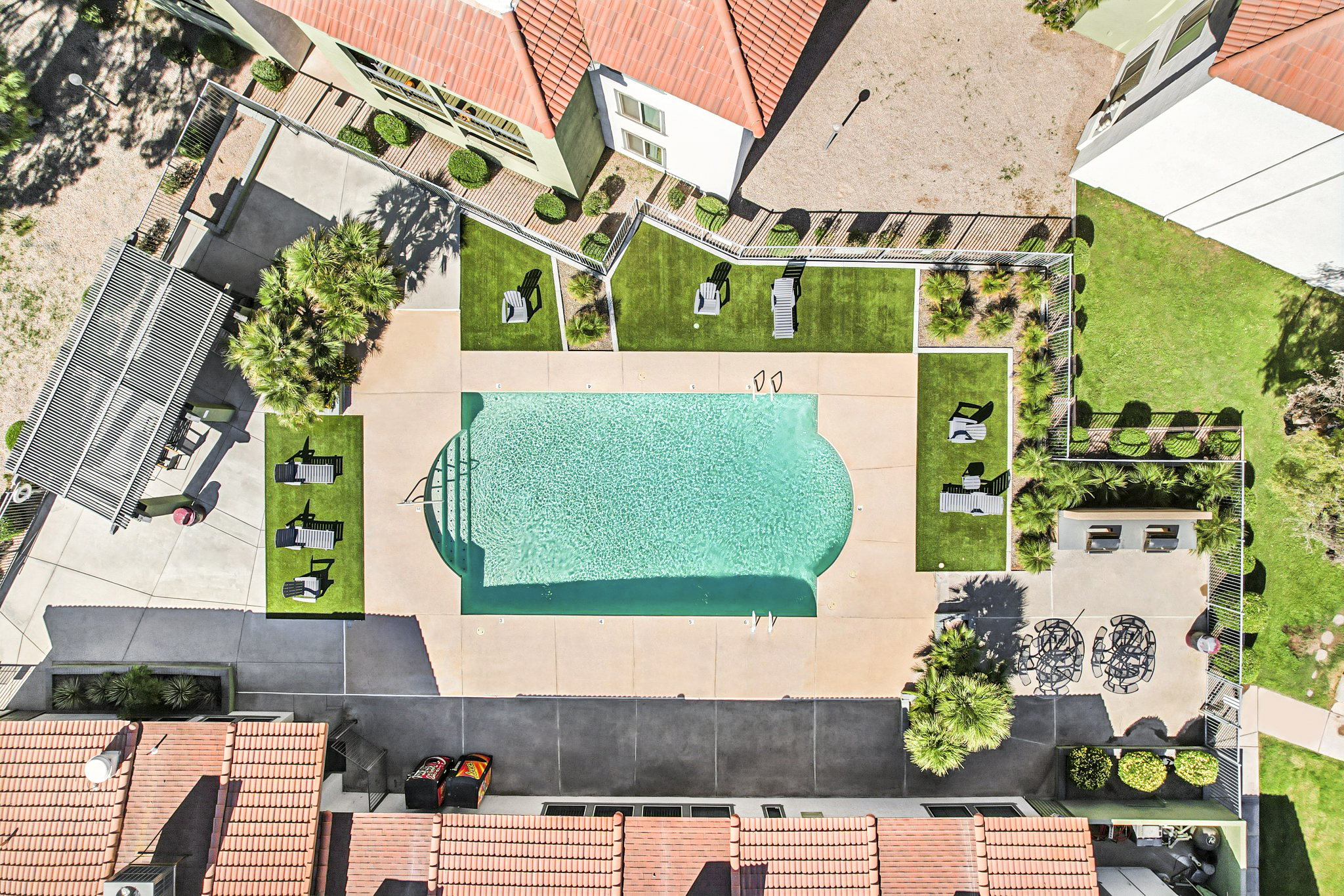 Aerial view of a swimming pool surrounded by lounge chairs and green grass, with nearby shaded seating areas and buildings with red roofs in the background. The pool area is well-maintained, showcasing clear water and inviting outdoor spaces.