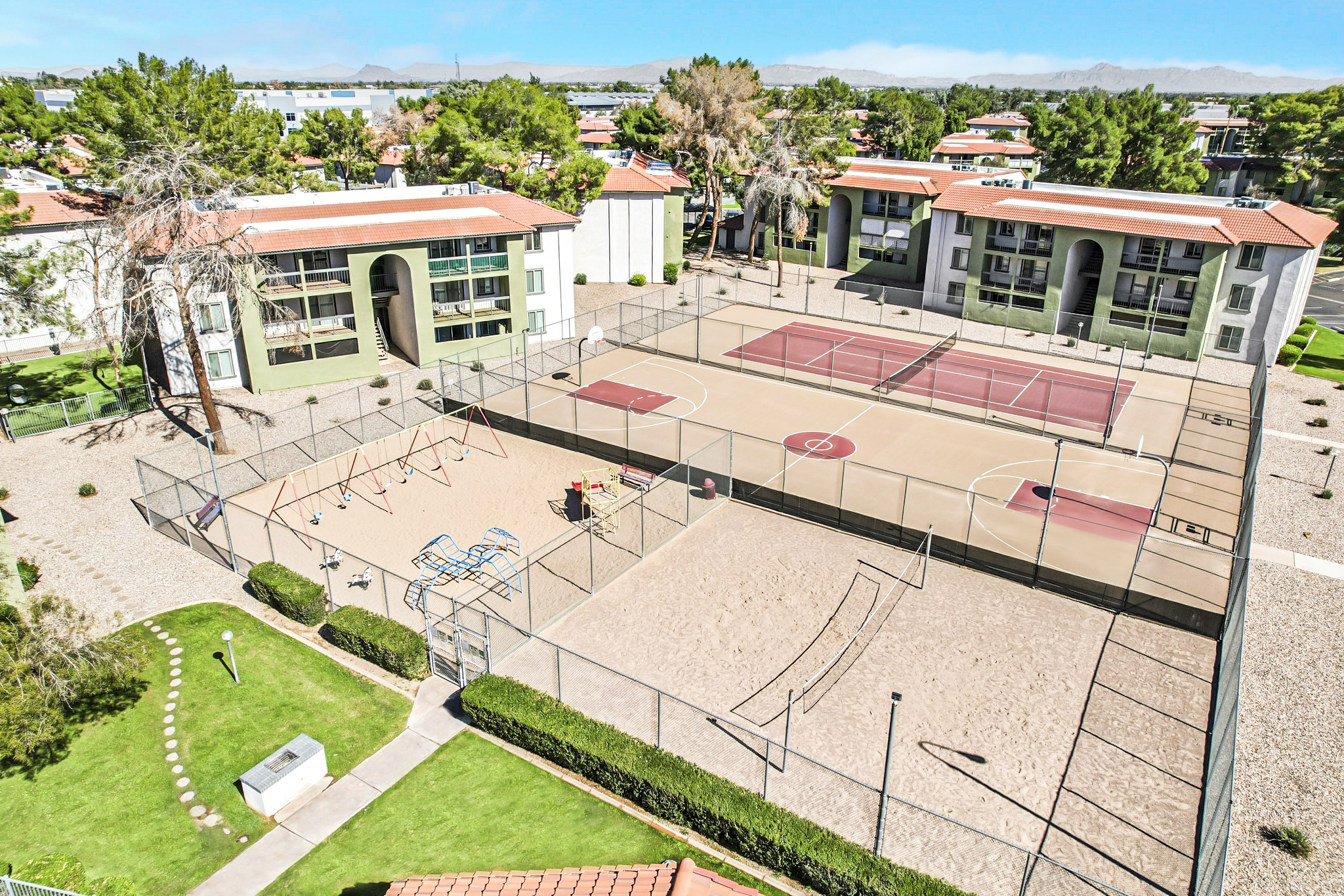Aerial view of a residential complex featuring tennis courts, a basketball court, and playground equipment. Surrounding the area are well-maintained grassy lawns and trees, with buildings in the background. The scene is bright and sunny, showcasing an inviting recreational space.