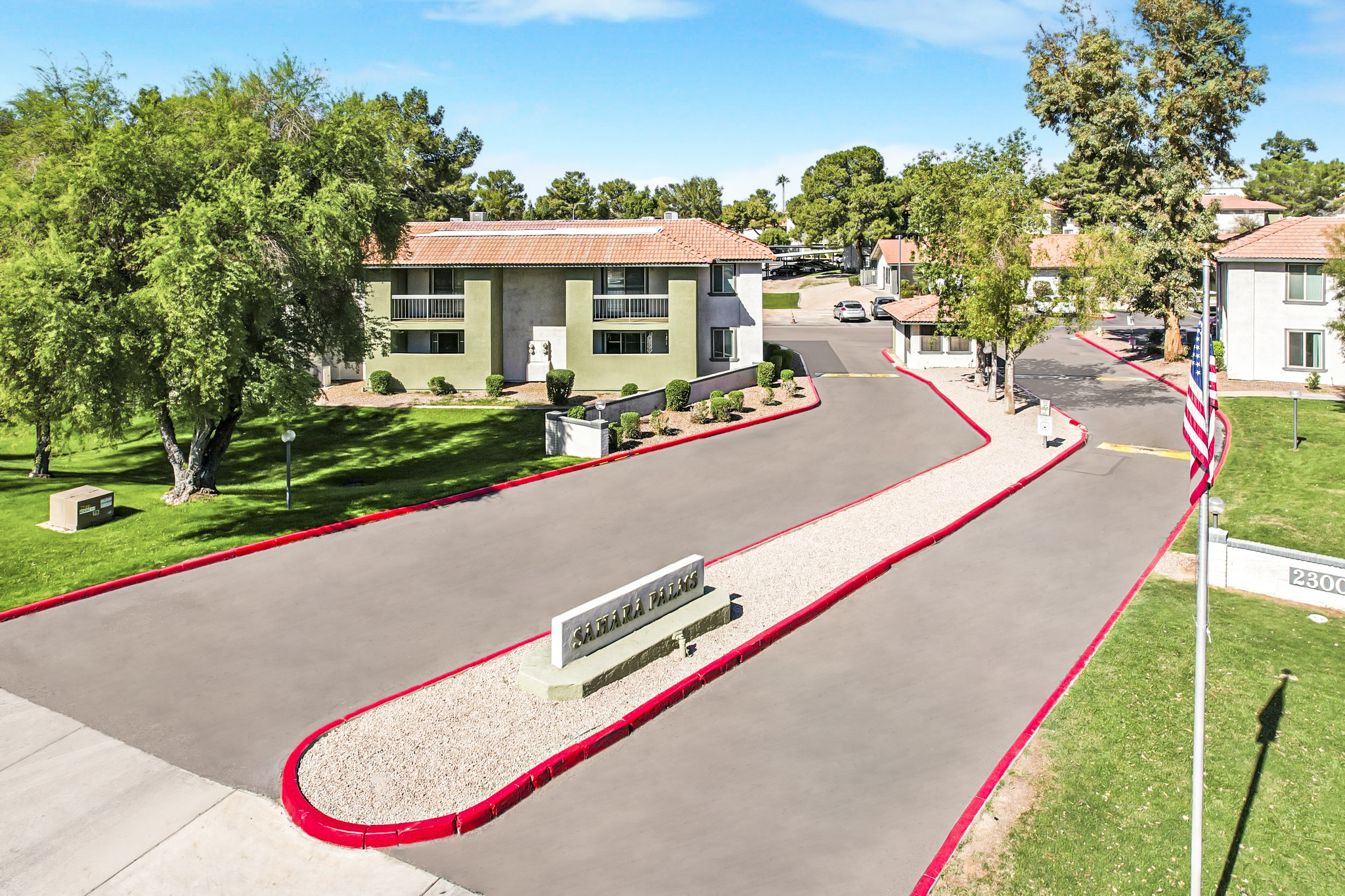 Aerial view of a residential complex with two buildings in the background, a landscaped entrance with a sign reading "Katerra Homes," and well-maintained greenery. The pathway is lined with red curbs and gravel, and there are trees nearby. An American flag is visible on the left side of the image.