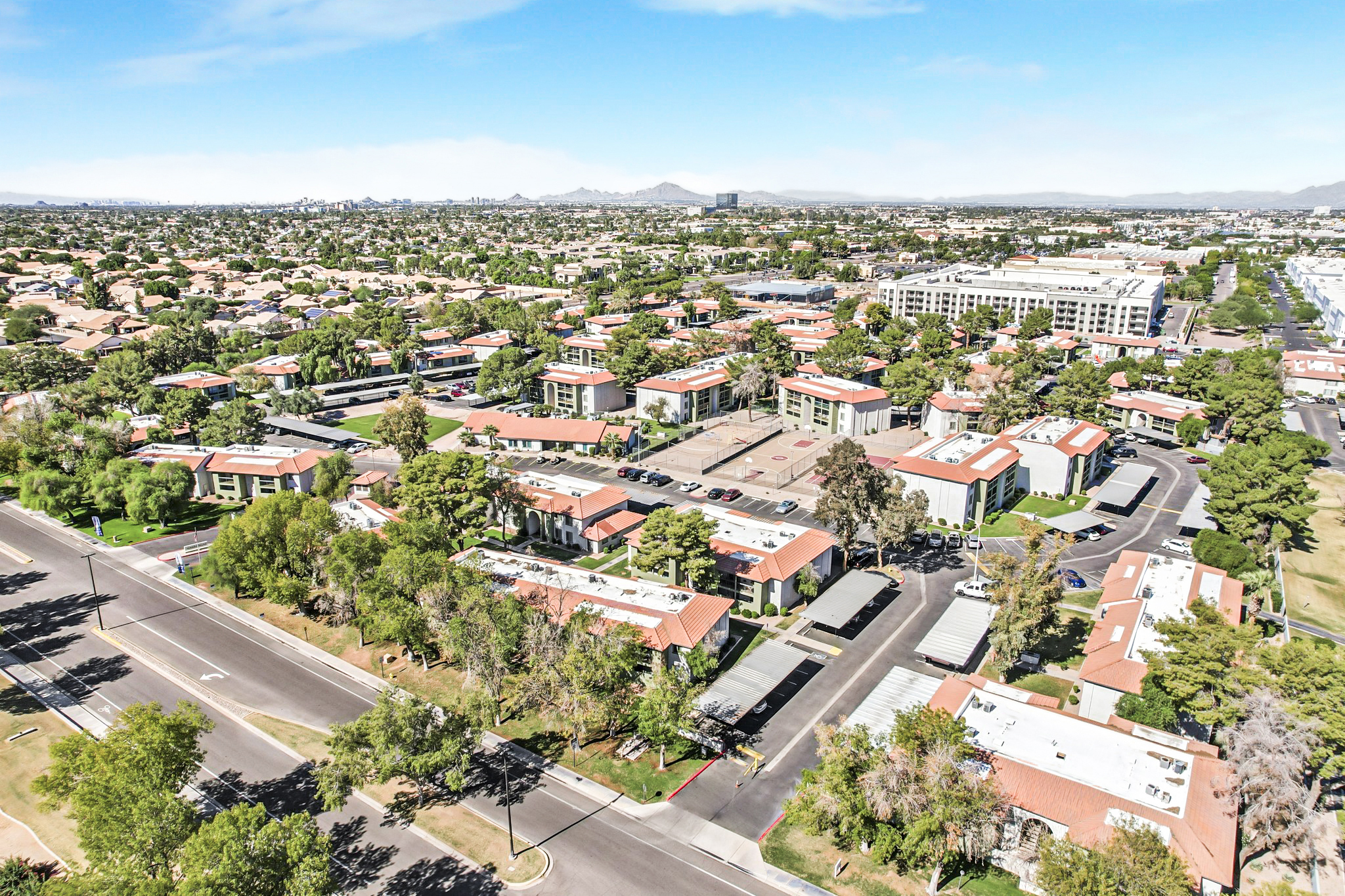 Aerial view of a suburban neighborhood featuring multiple residential buildings with red roofs, landscaped green areas, and nearby roadways. The landscape shows a mix of apartment complexes, trees, and open spaces under a clear blue sky. In the background, mountains and additional city structures are visible.