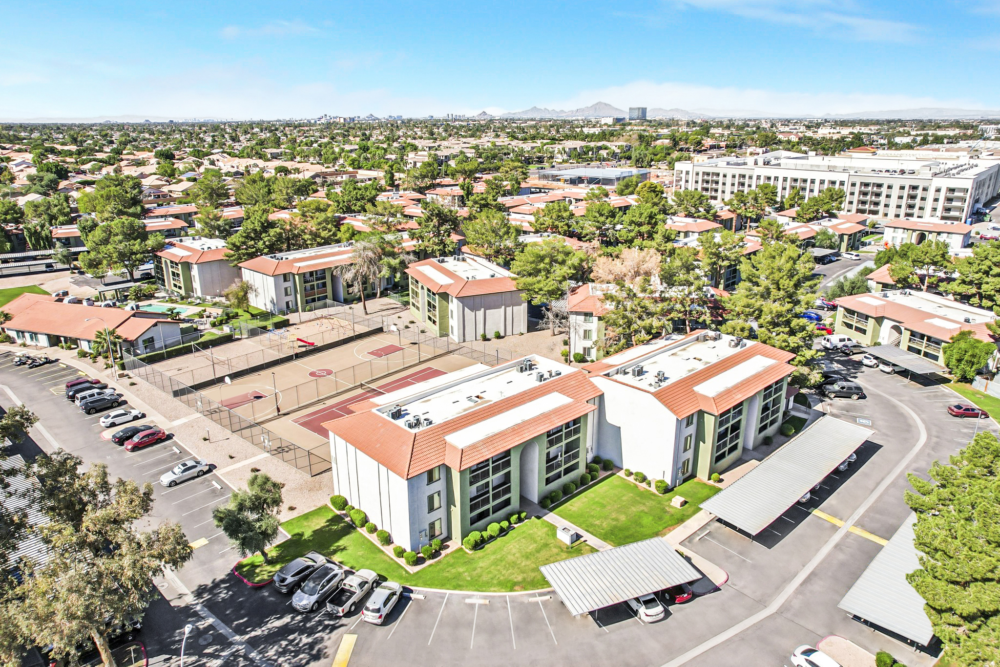 Aerial view of a residential area featuring several multi-story apartment buildings with red roofs. The complex includes a parking lot, basketball courts, and green landscaping. In the background, there are more buildings and a mountain range under a clear sky.
