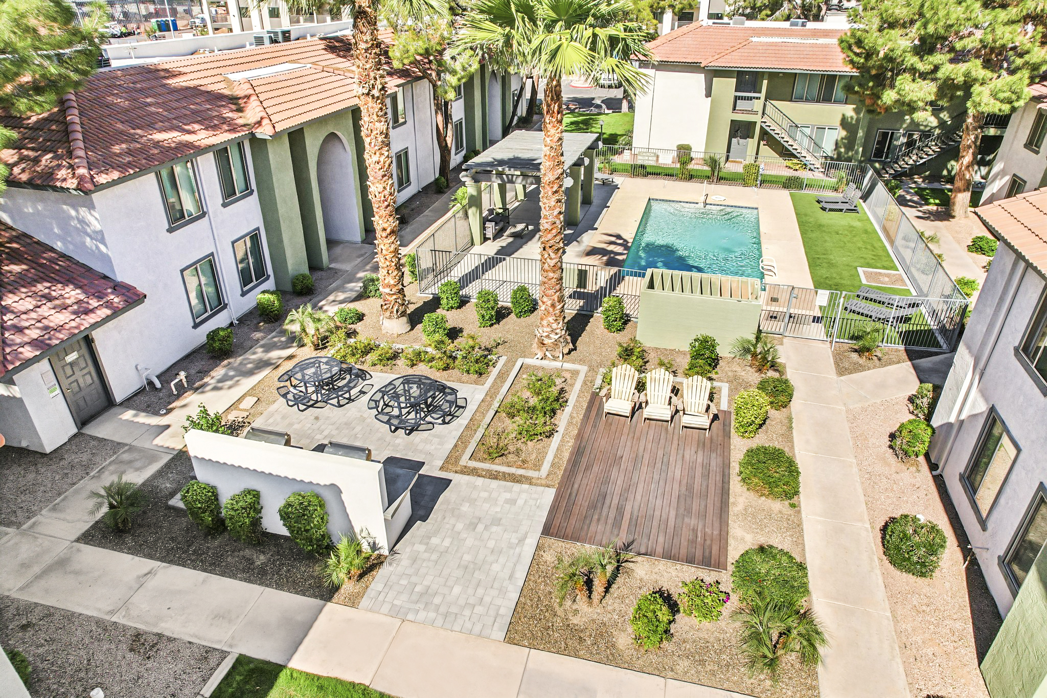 Aerial view of a modern residential complex featuring a pool, palm trees, and landscaped areas. There are lounge chairs and outdoor seating arrangements, with pathways and greenery surrounding the buildings. The scene conveys a relaxed and inviting atmosphere for residents.