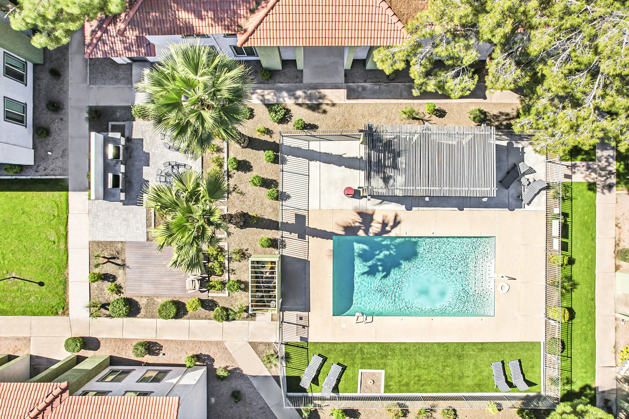 Aerial view of a residential area featuring a swimming pool surrounded by lounge chairs and palm trees. Adjacent to the pool, there is a covered seating area and a paved patio. The surrounding landscaping includes neatly trimmed grass and shrubs, with pathways connecting the buildings.