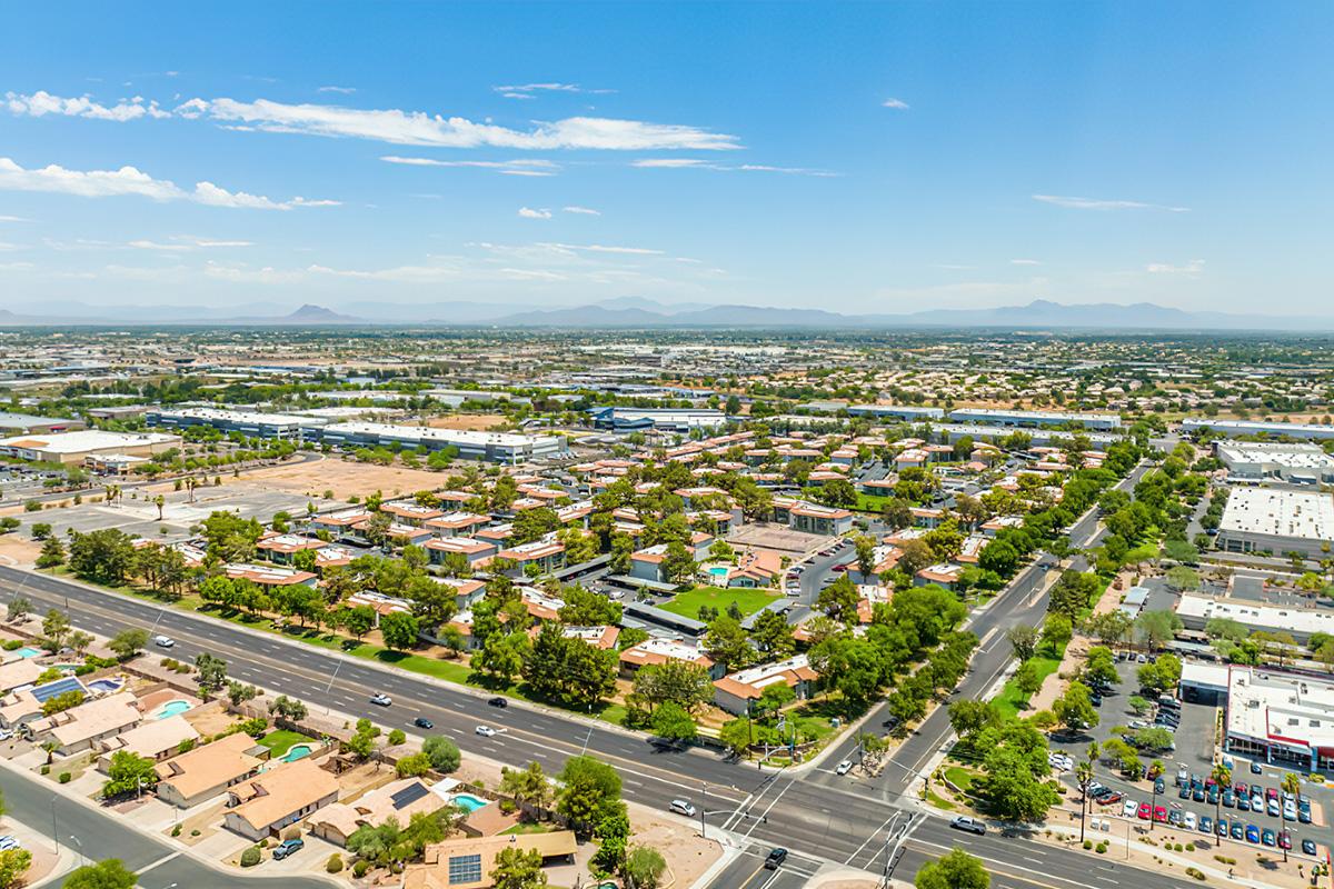 Aerial view of a residential area featuring a mix of single-family homes and apartment communityes, with tree-lined streets and landscaped yards. In the distance, mountains are visible under a clear blue sky, while roads are bustling with cars. The scene reflects a suburban community surrounded by commercial properties.