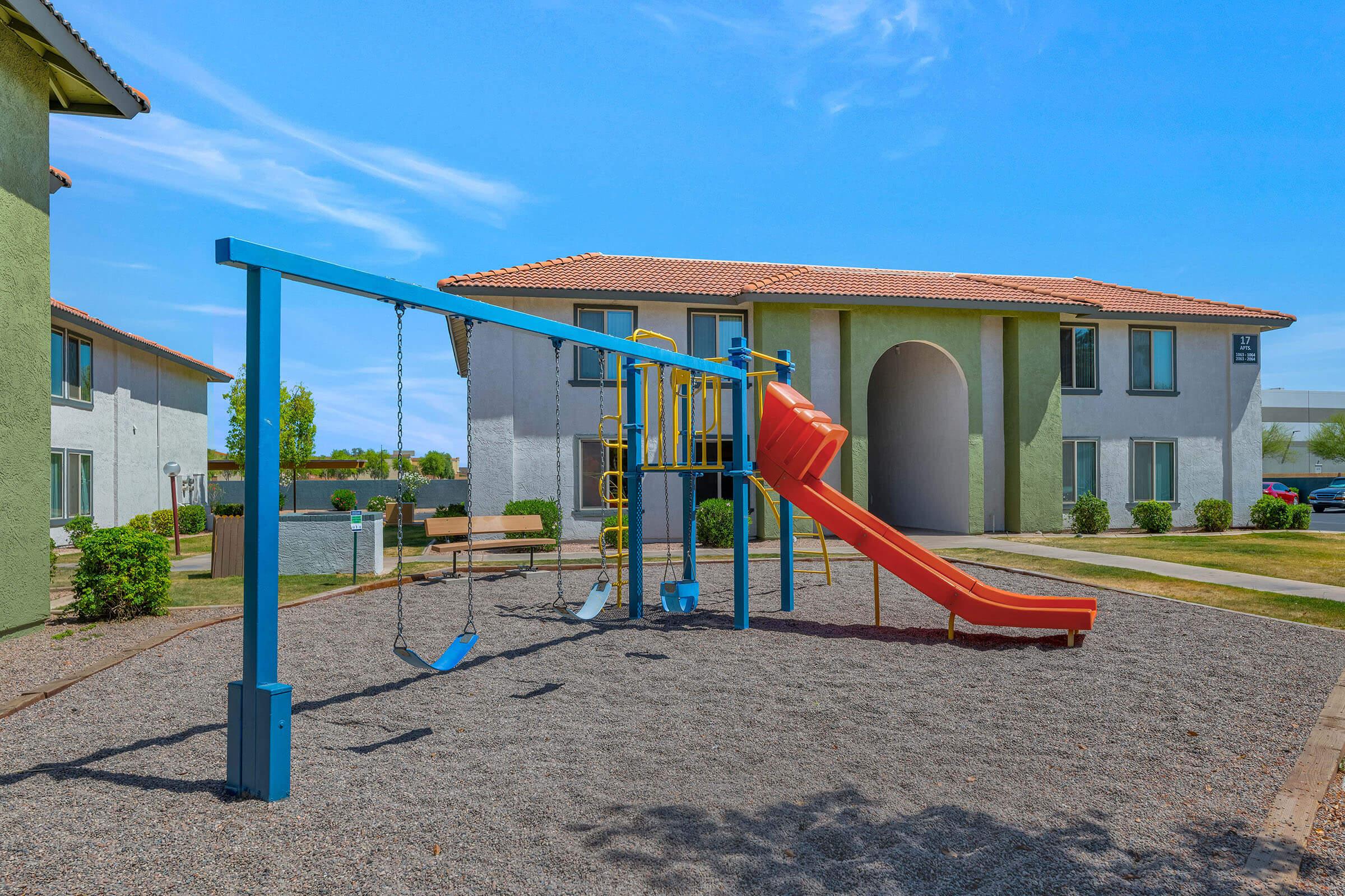 A colorful playground featuring swings and a slide, located in a gravel area in front of a residential building. The sky is clear with a few clouds, and there are landscaped shrubs surrounding the playground. The building has a light green exterior with terracotta roof tiles.