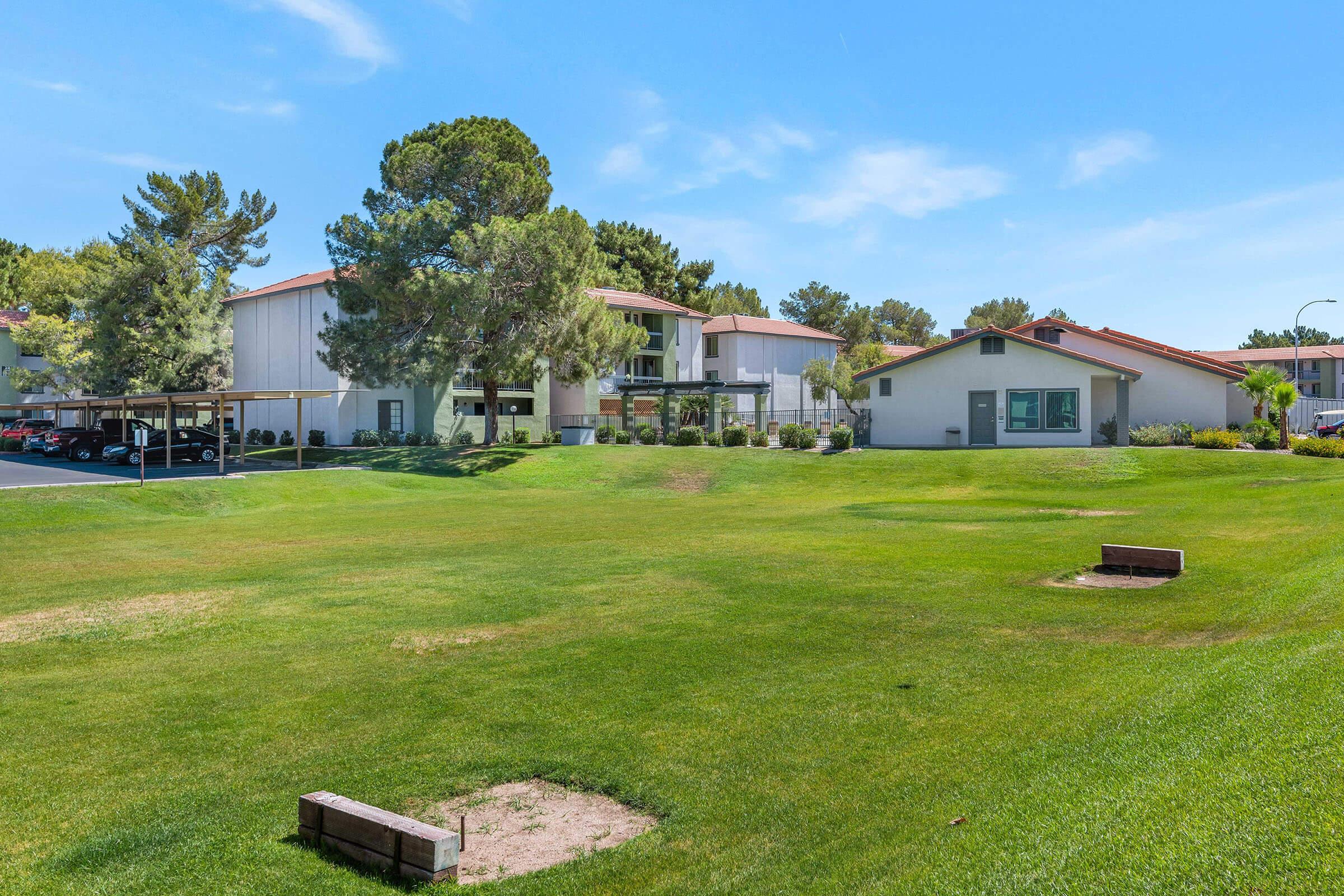 A well-maintained lawn with two stone benches and a backdrop of residential buildings surrounded by trees. The sky is clear and blue, emphasizing the greenery and inviting outdoor space. Parking can be seen to the left, providing convenient access to the buildings.