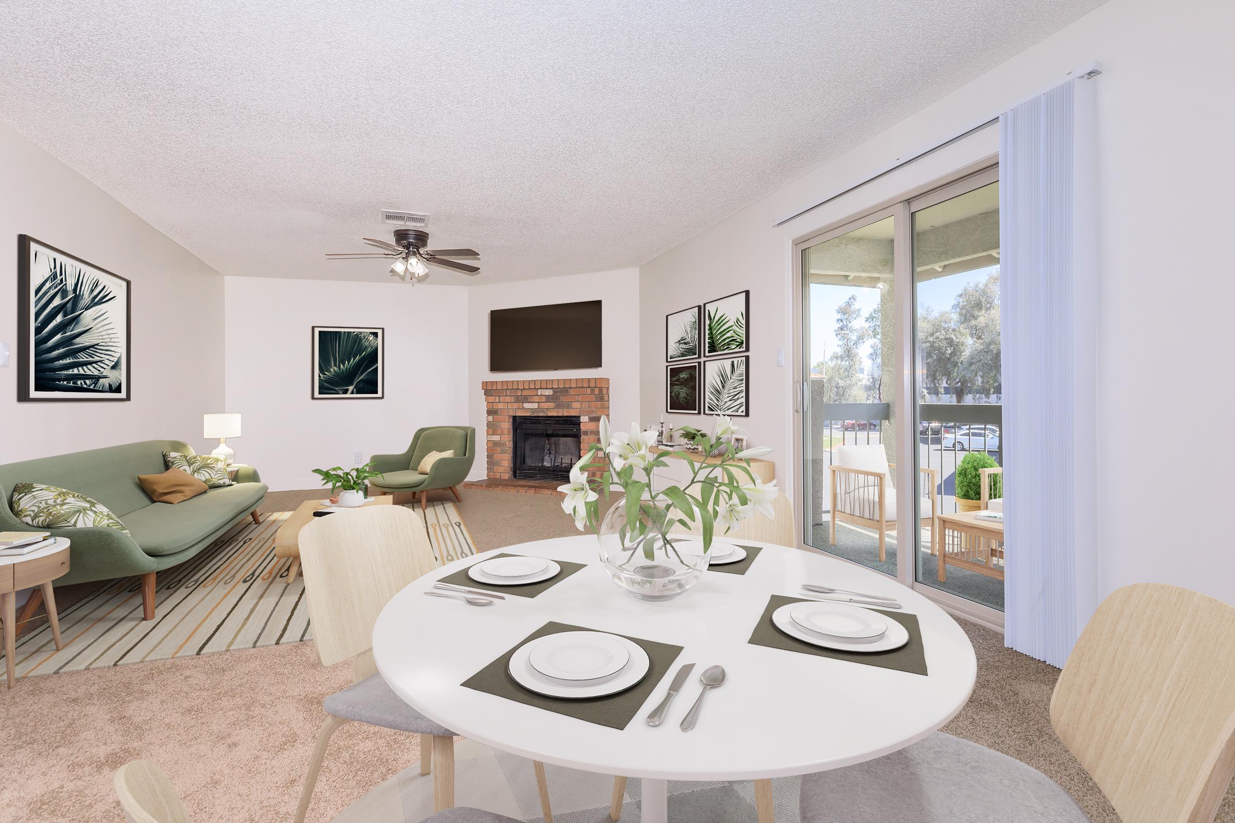 A modern living area featuring a round dining table set with plates and cutlery. In the background, there is a green sofa, a fireplace, and framed botanical artwork on the walls. Large windows with sheer curtains let in natural light, revealing a view of the outdoors and patio furniture.